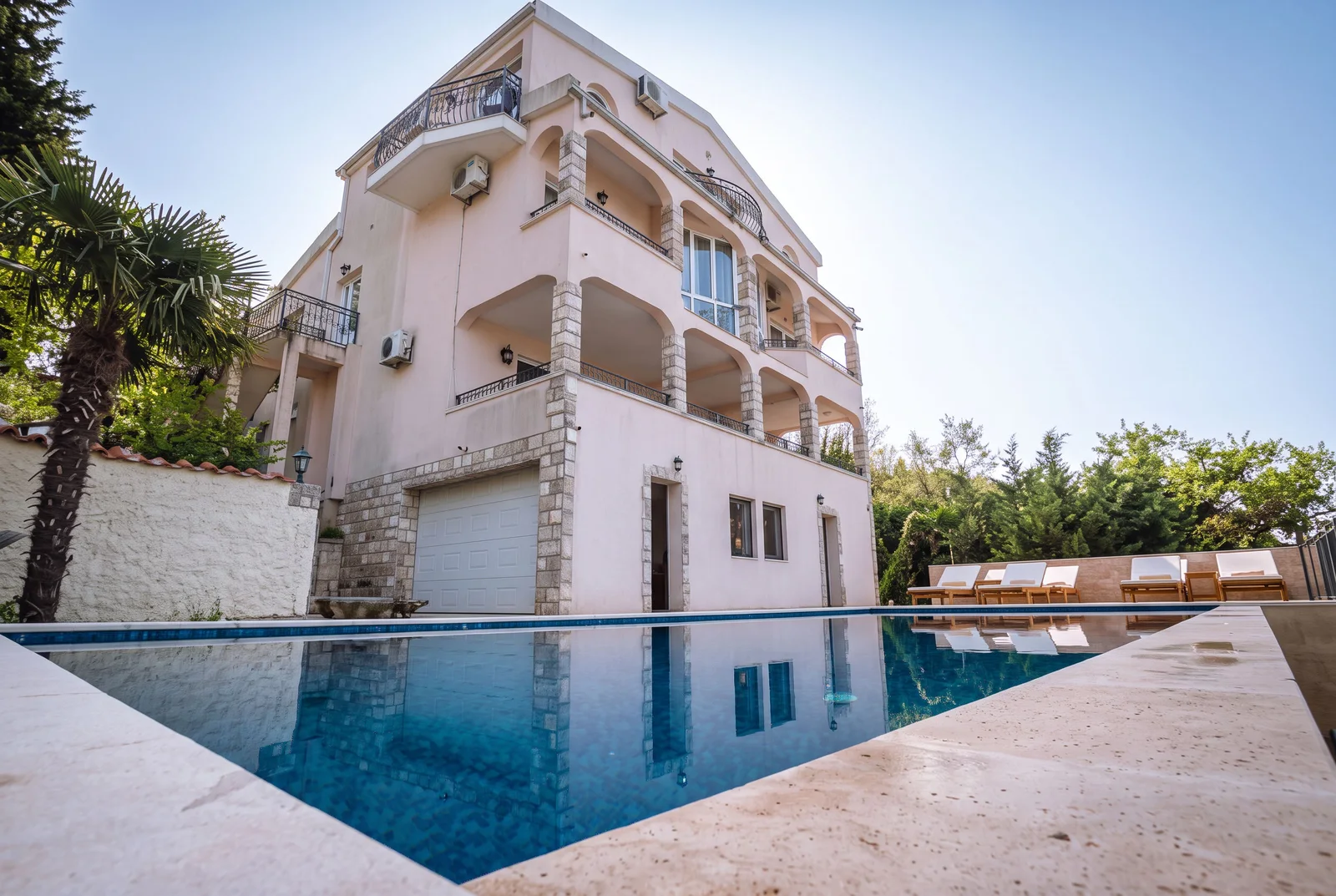 Swimming pool in foreground, pink multi-story house with balconies and garage in background.