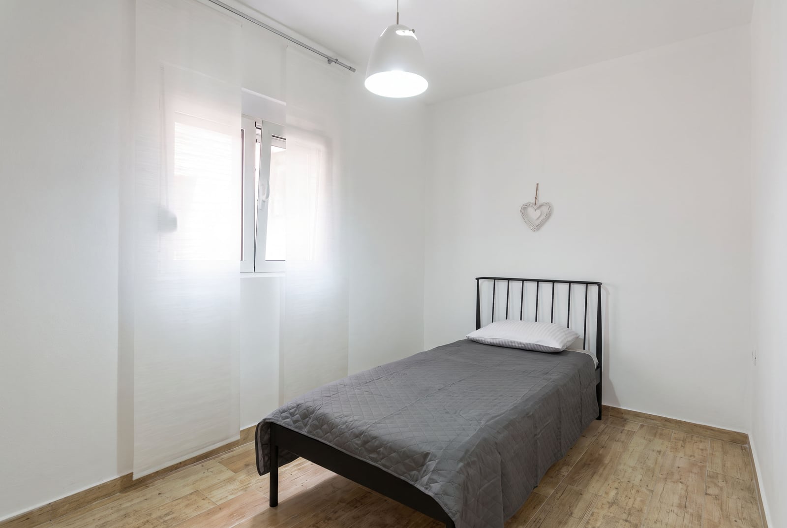Small bedroom featuring a single bed with a black metal frame, gray quilted cover, white pendant light, and sheer window curtains.