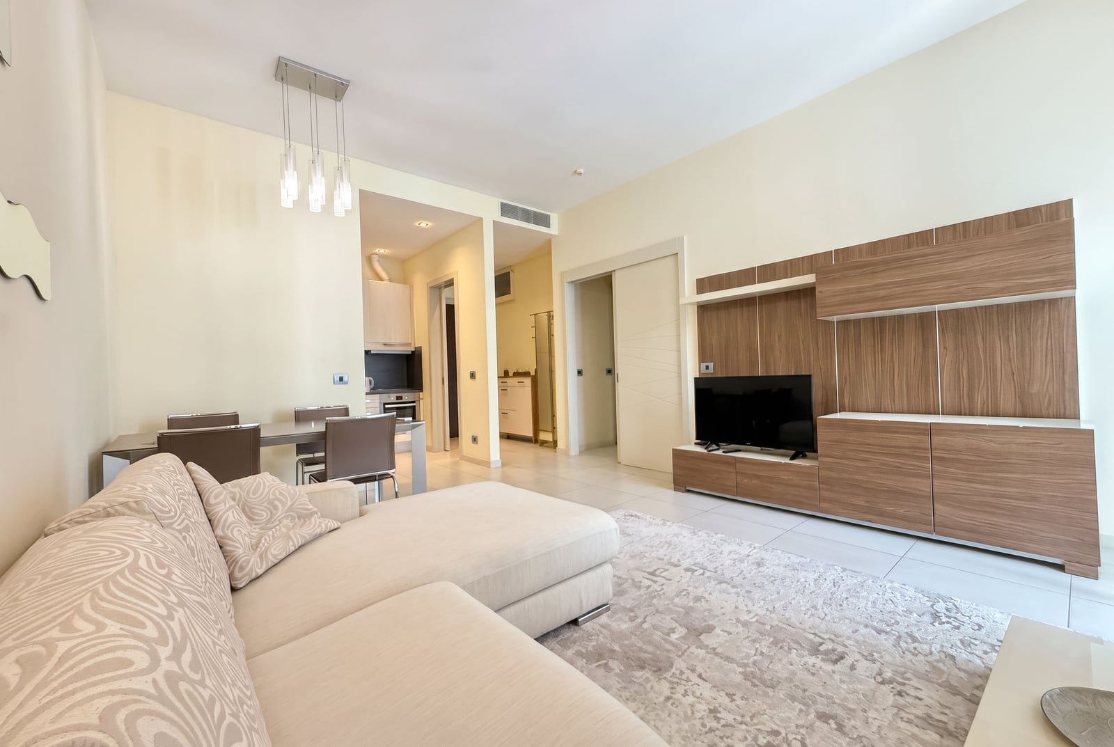 Bright living room with a beige sofa, wood TV wall unit, and dining area visible in the background.