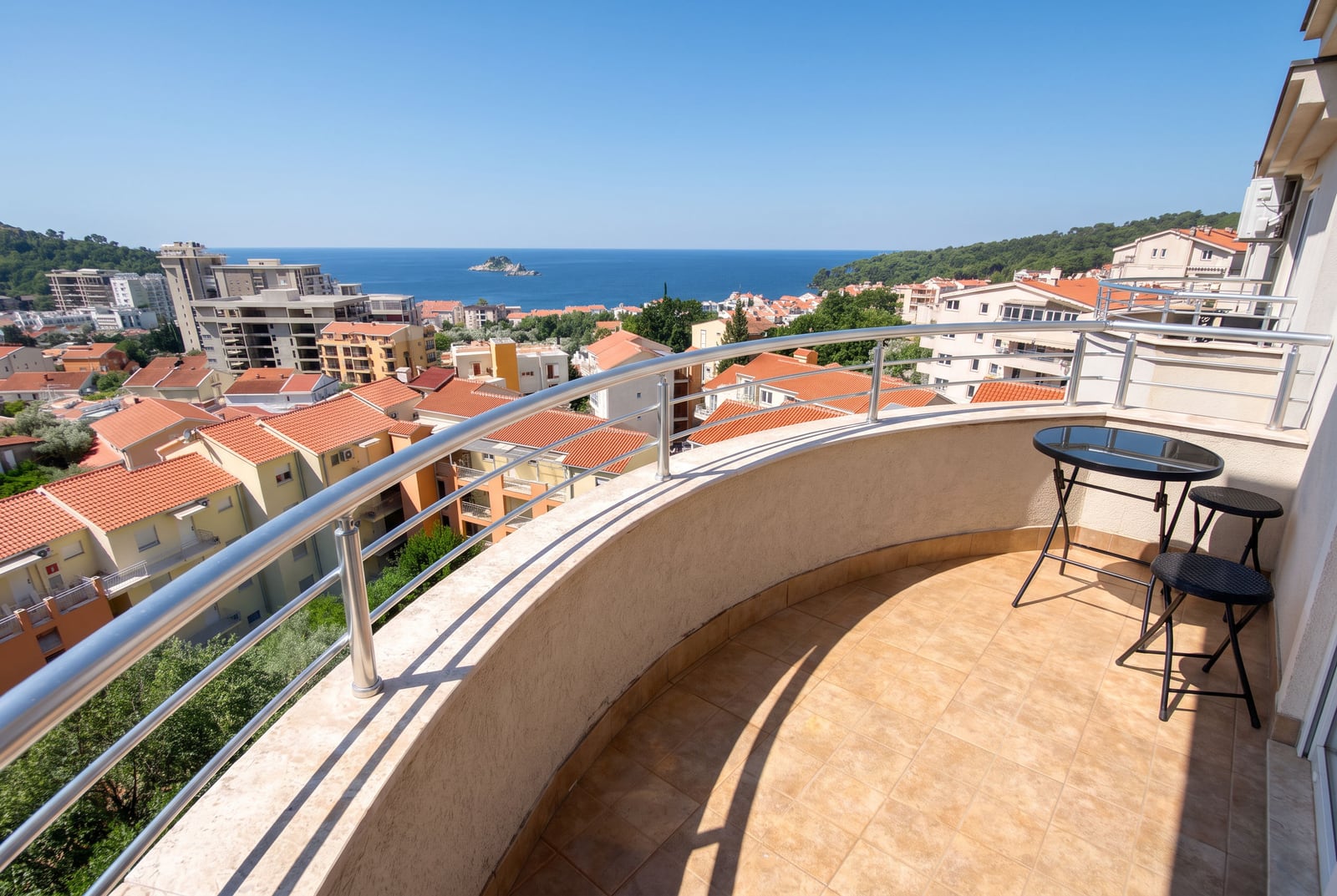 Curved balcony with metal railing, black table, and view of city rooftops and the sea.