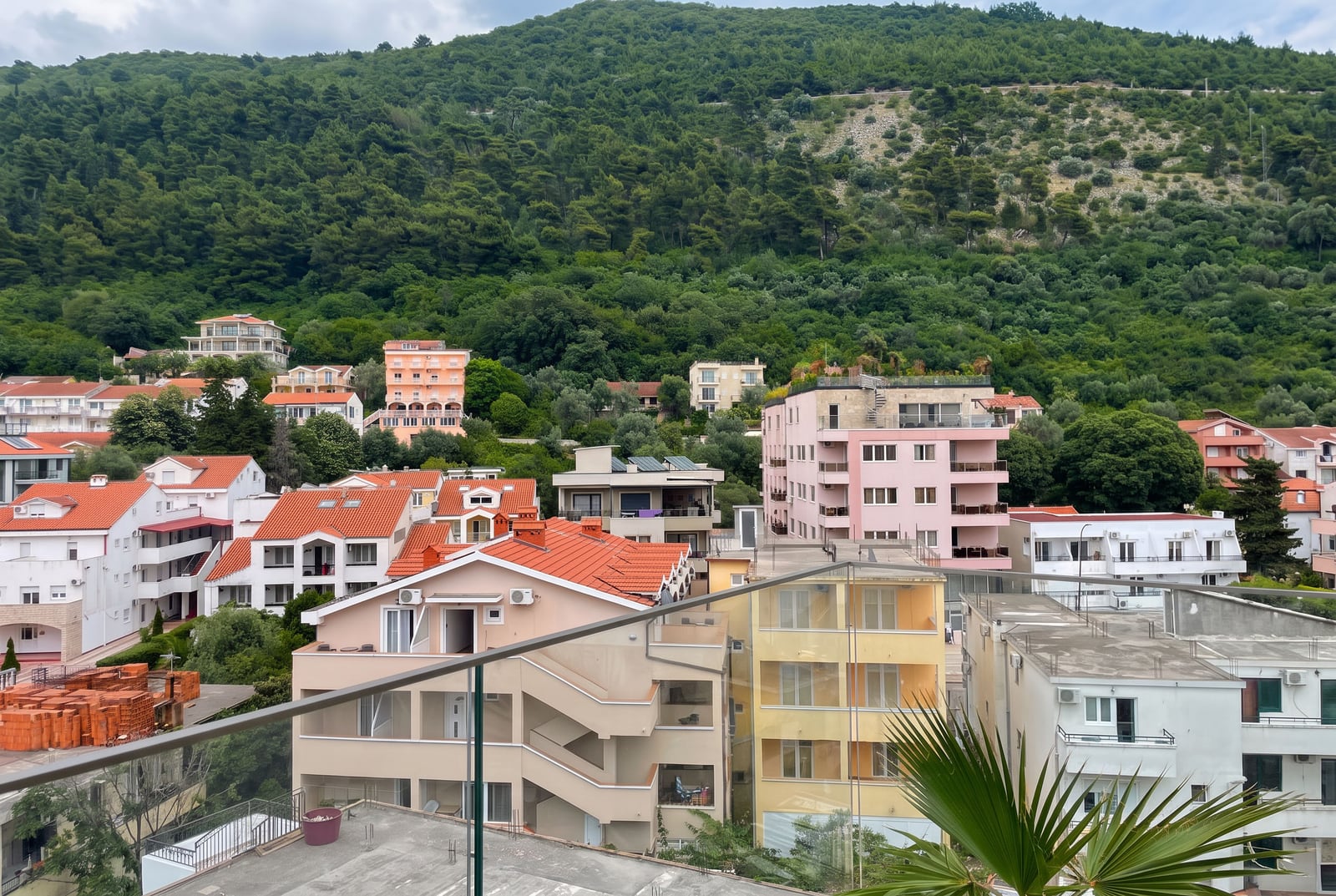 View from a balcony showing residential buildings with red roofs and a green forested hill.