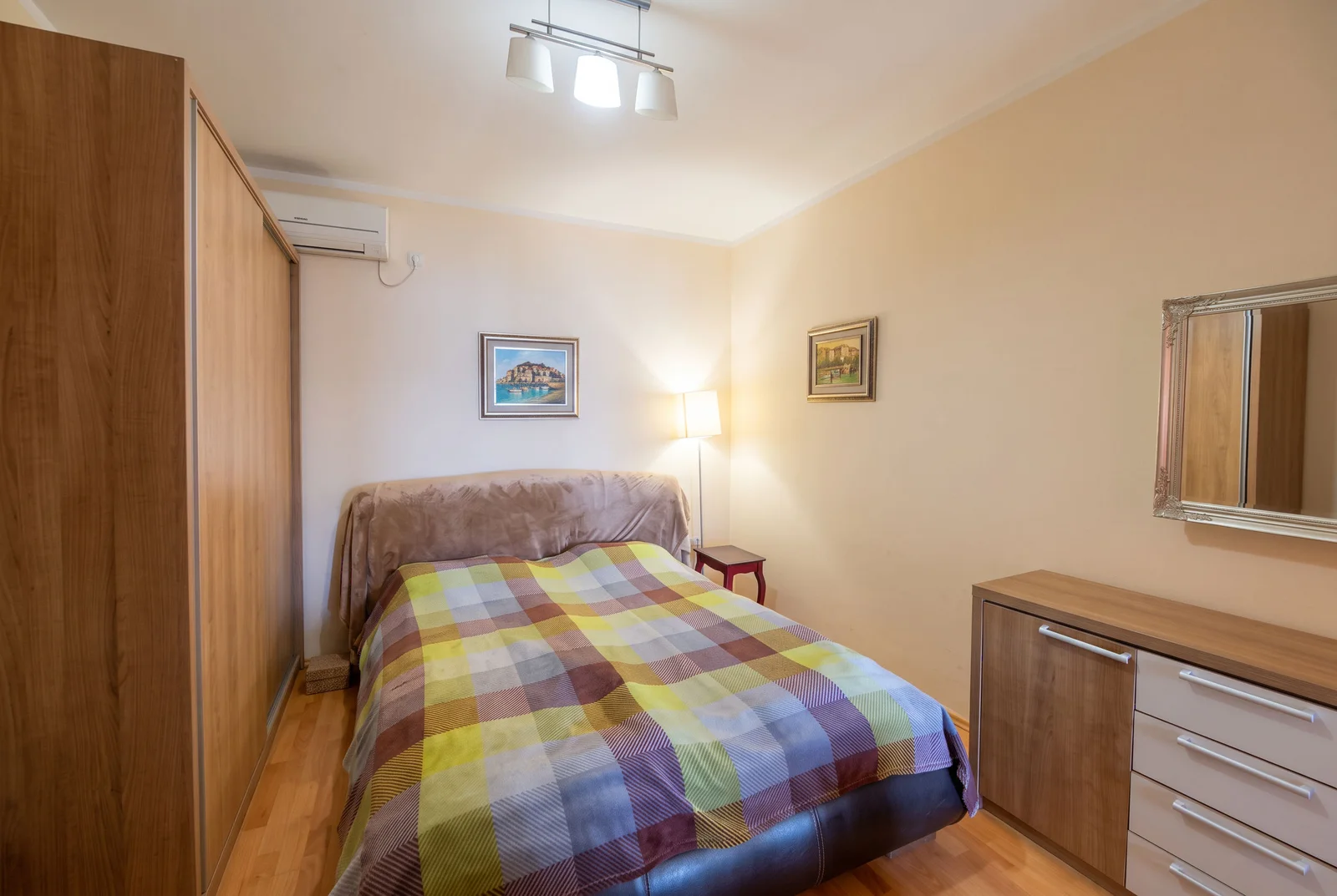 Bedroom featuring a double bed with plaid cover, wooden wardrobe, dresser with mirror, and wall art.