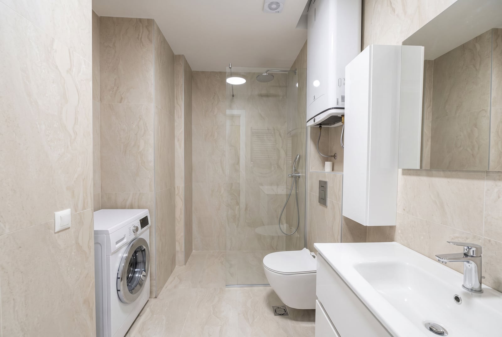 Modern bathroom featuring a glass shower, washing machine, wall-mounted toilet, and sink on beige tiles.