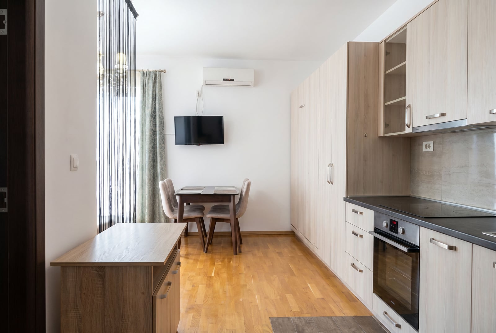 Kitchen-living room in an apartment in Budva, Montenegro: light-colored kitchen set, dining area, parquet flooring, and built-in appliances.