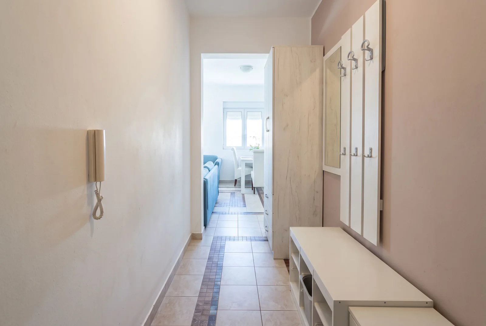Entryway hallway featuring a white storage unit, bench, wall phone, and mirror leading to a living area.