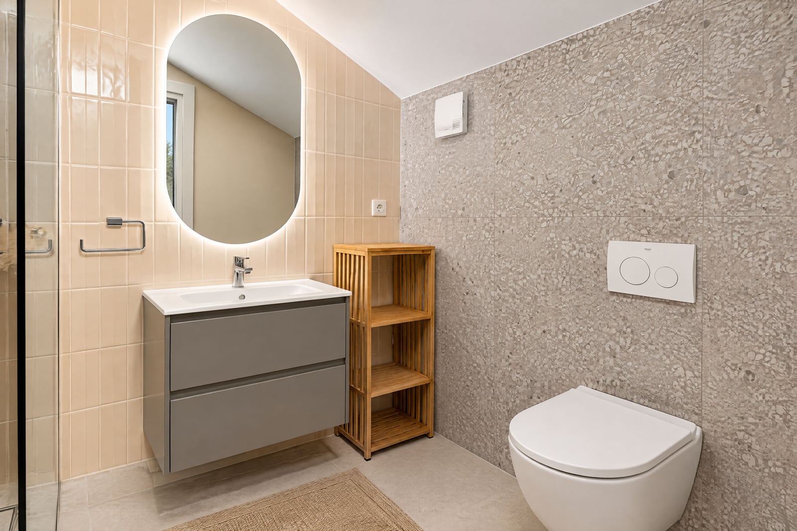 Modern bathroom featuring a grey vanity, round backlit mirror, wall-mounted toilet, and mixed beige tile with terrazzo finish.