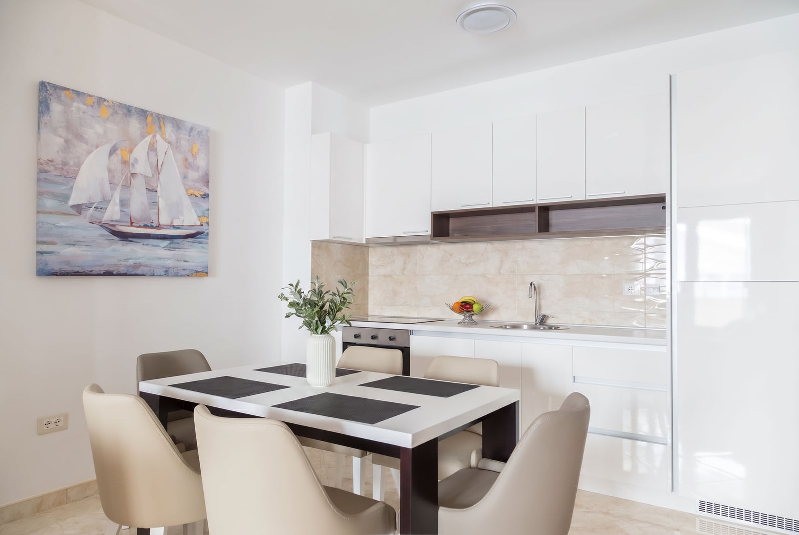 Modern kitchen-dining room in an apartment in Bečići, Montenegro: white furniture, dining table, and a painting of a sailboat.