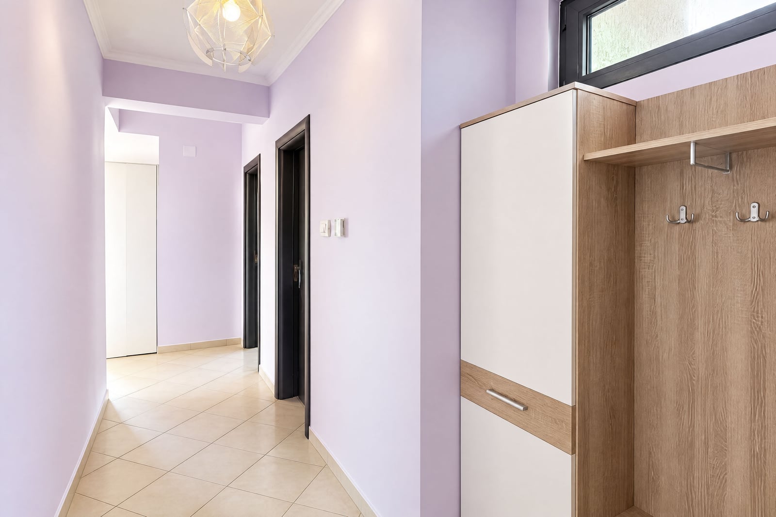 Hallway with lavender walls, tiled floor, and a wooden wardrobe unit with coat hooks on the right side.
