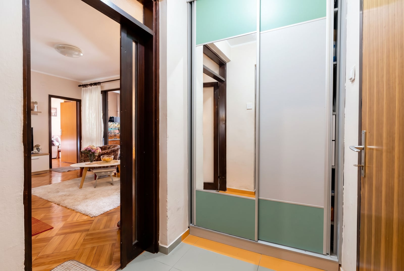 Entrance hall with a mirrored sliding-door wardrobe in an apartment for sale in Budva, Montenegro, overlooking a cozy living room with parquet flooring.