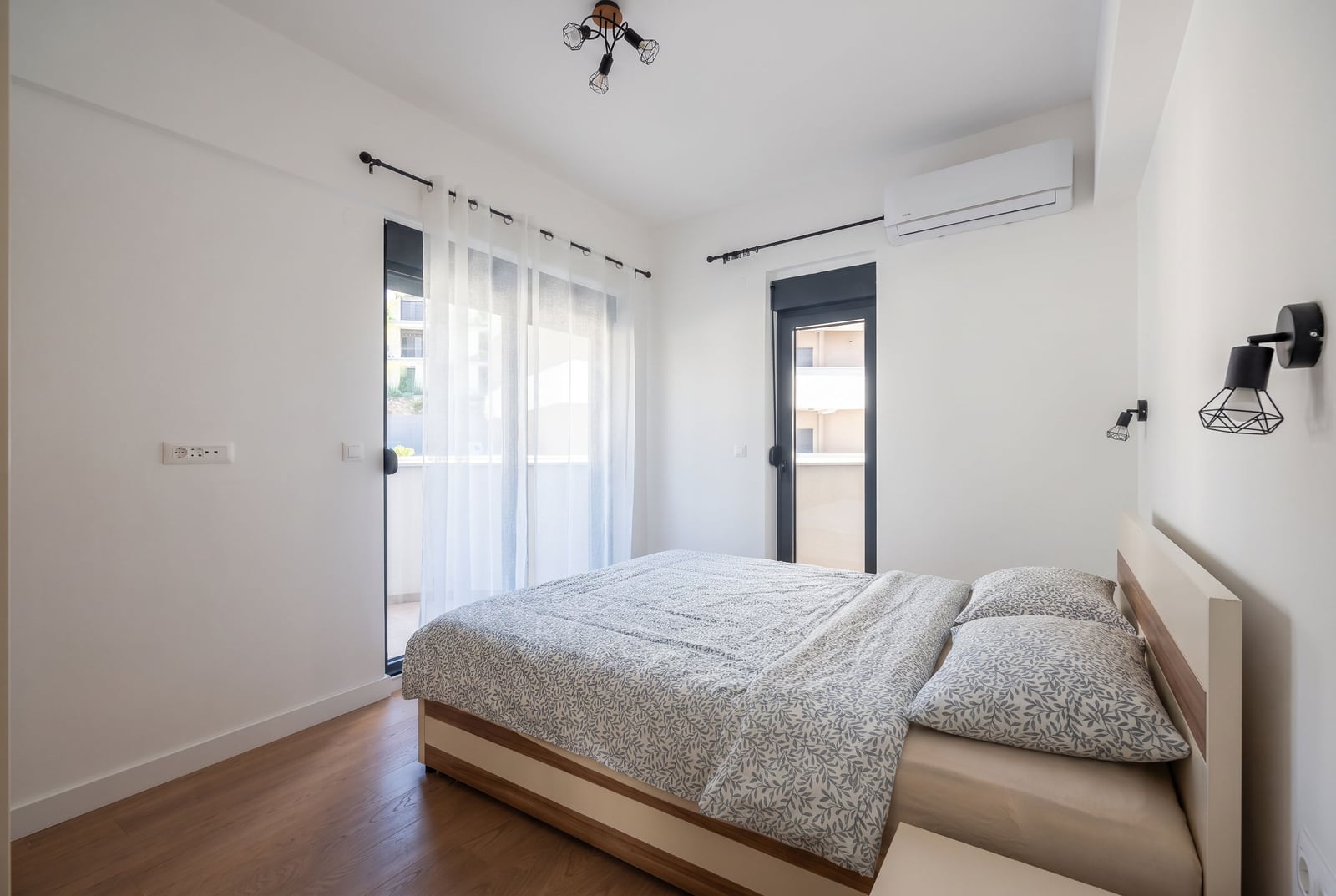 Bright bedroom with a double bed, wood flooring, AC unit, and balcony accessible via glass doors with sheer curtains.