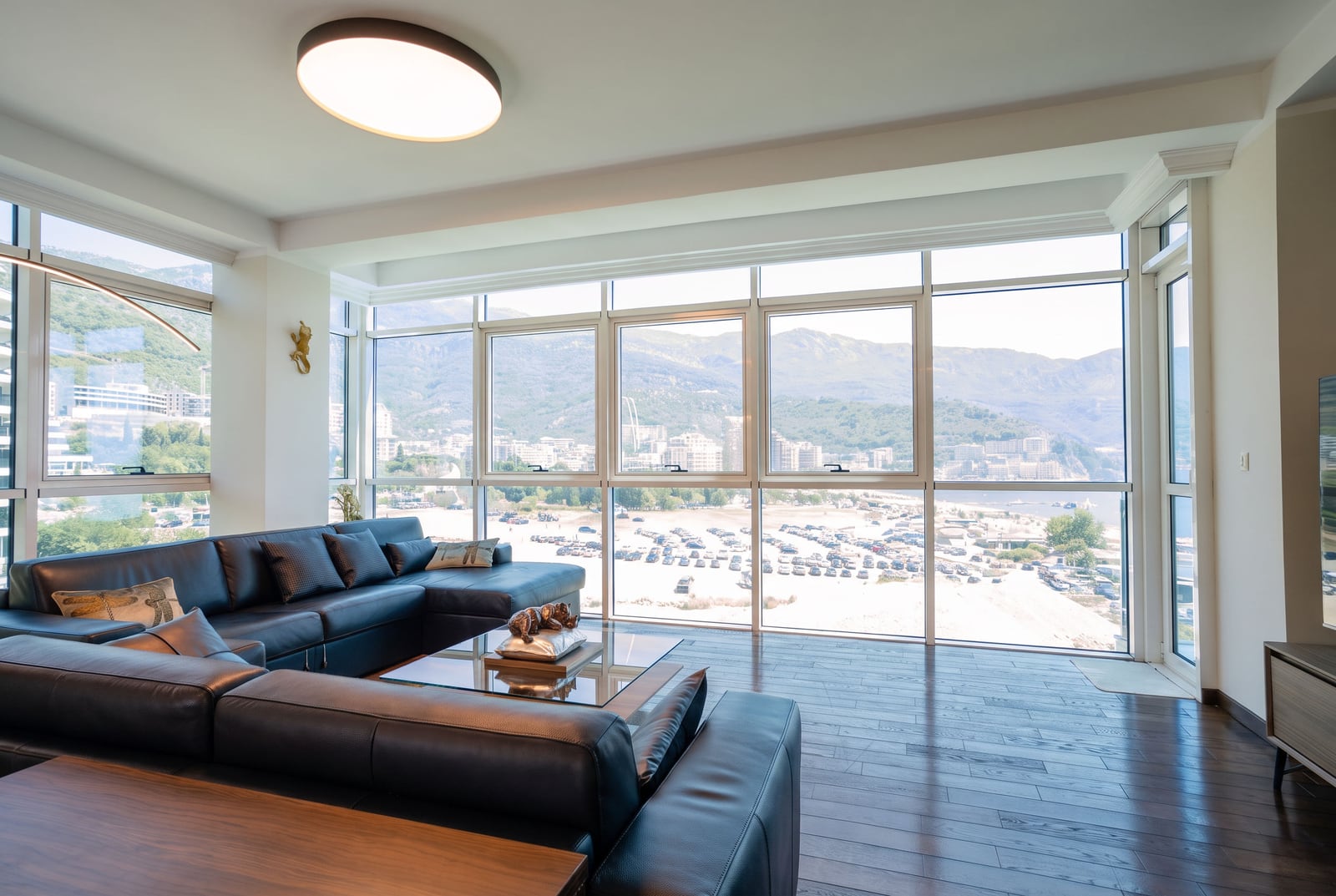 Bright living room with a large black leather sofa and panoramic windows overlooking mountains and a beach.