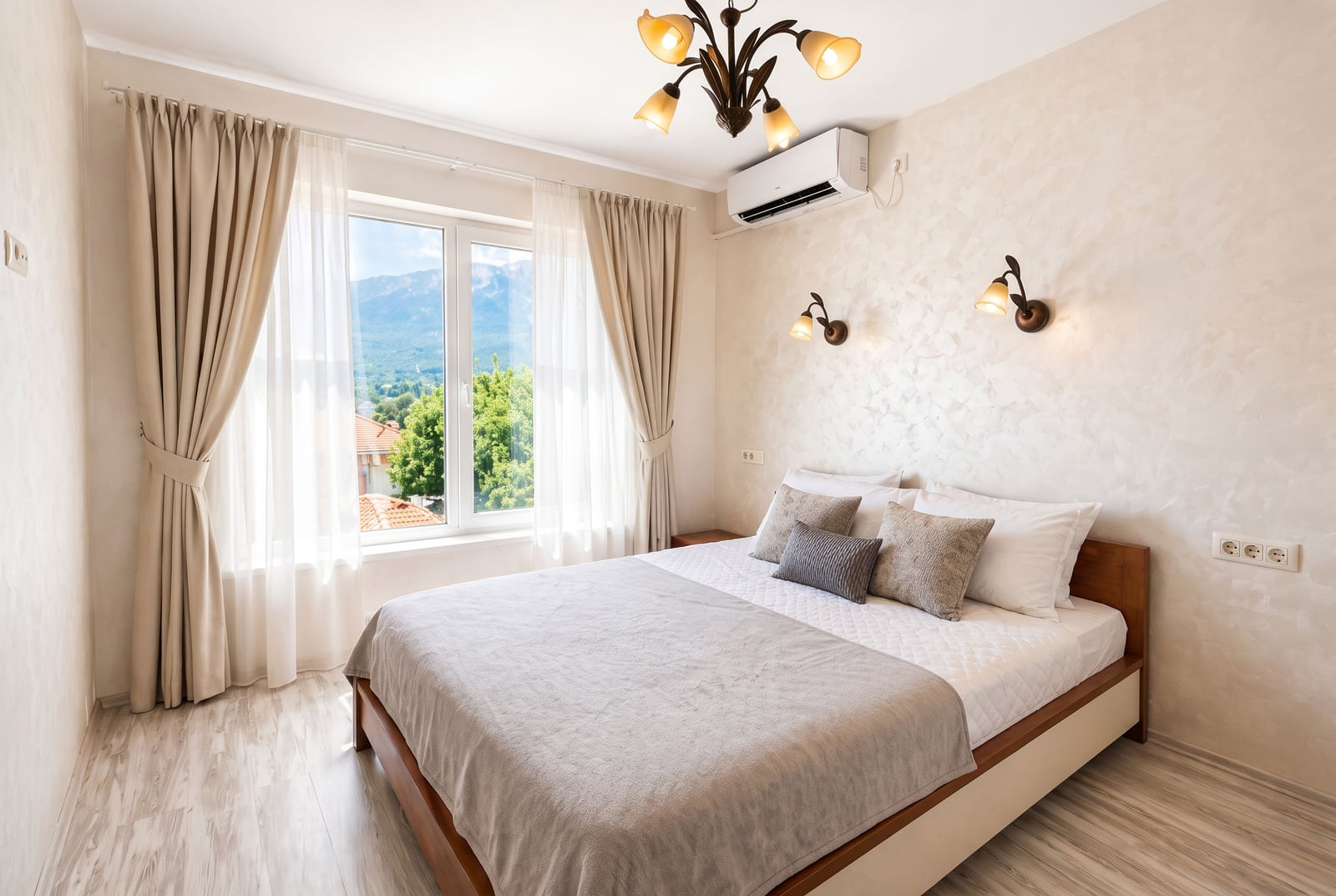 Bright bedroom with a wooden bed, beige curtains, wall sconces, and a mountain view through the window.