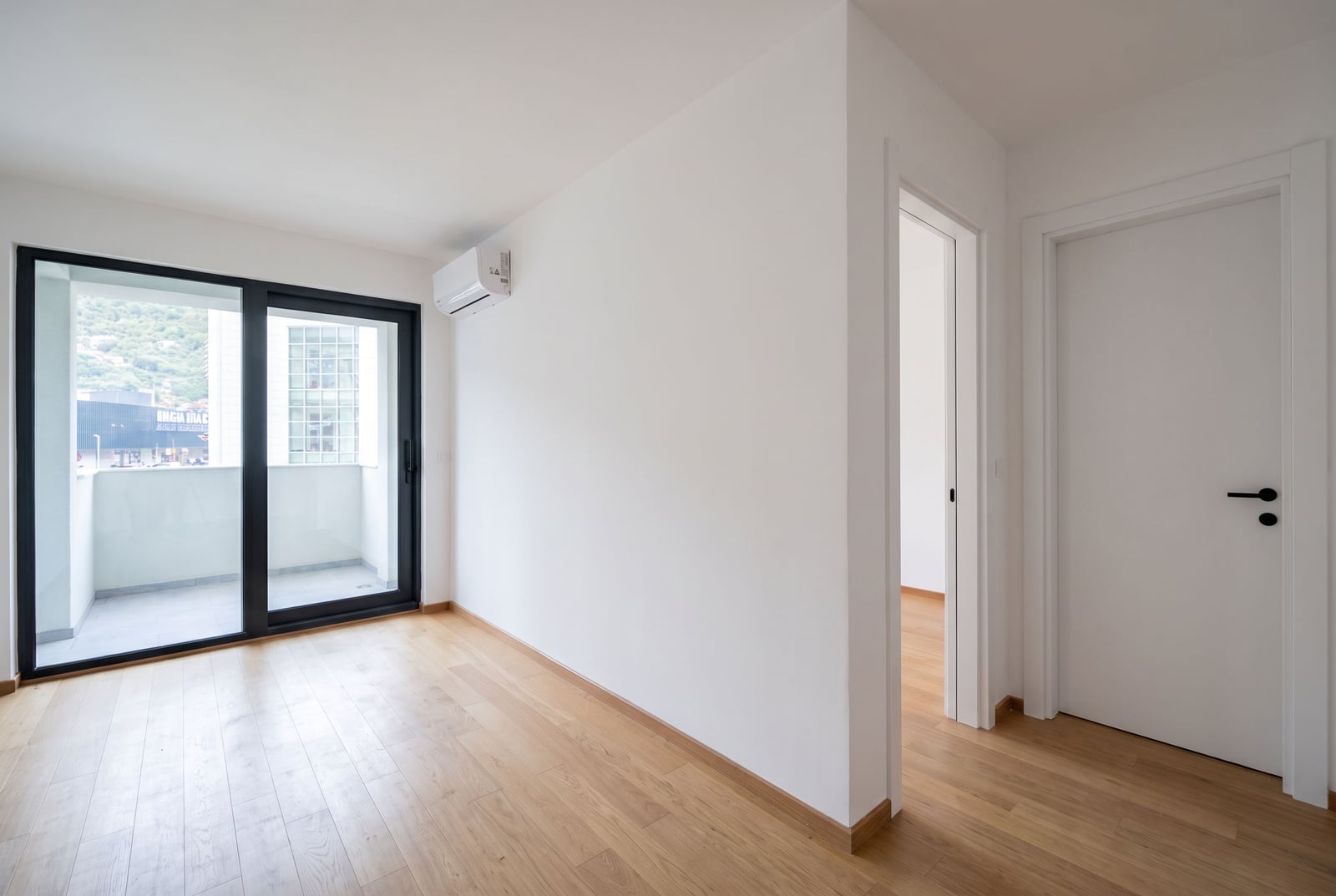 Bright empty living room with panoramic window, parquet flooring, and access to a balcony in a new apartment in Budva, Montenegro