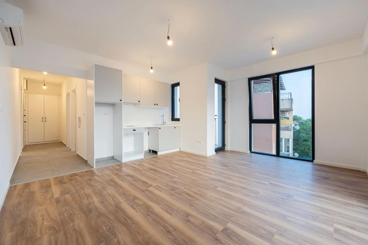 Open living area with wood flooring, white kitchenette, large window, and hallway visible.
