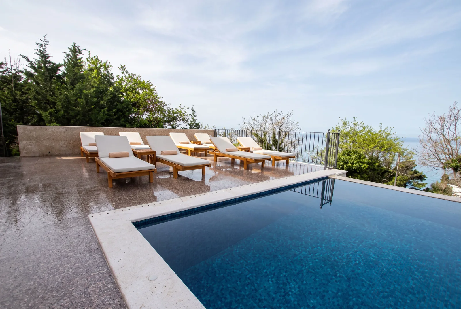 Infinity pool with wooden lounge chairs on a terrace overlooking the sea and trees.