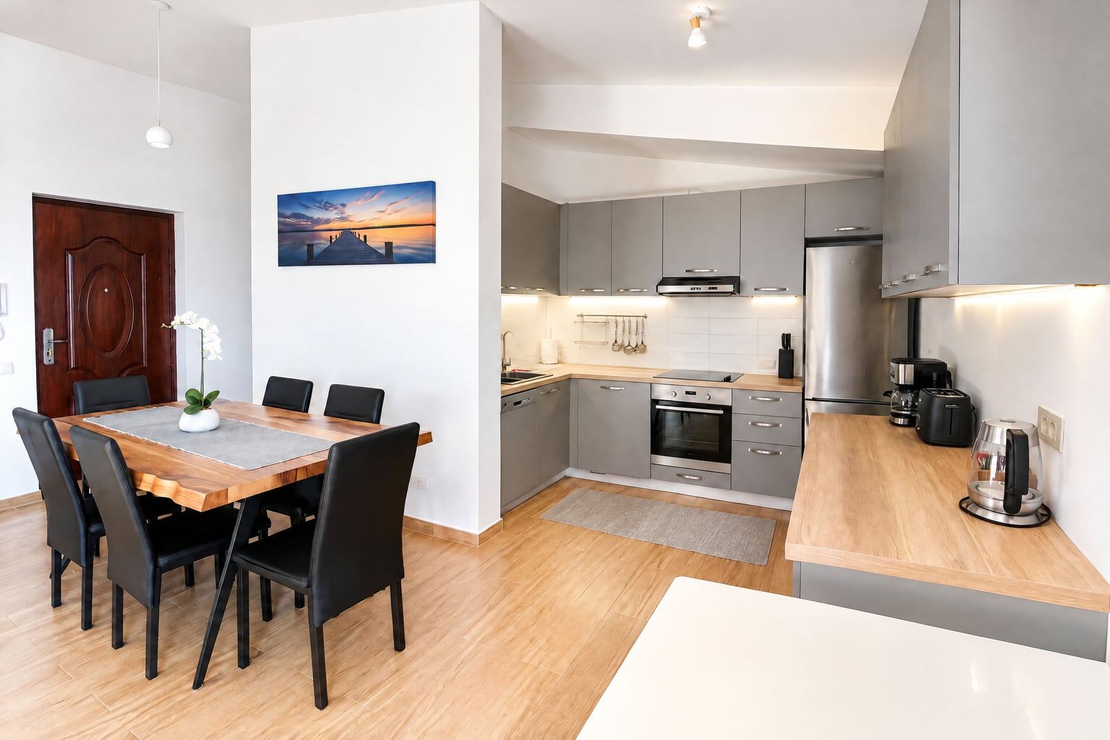 Modern kitchen with grey cabinets, wooden dining table, black chairs, and built-in appliances like oven and fridge.