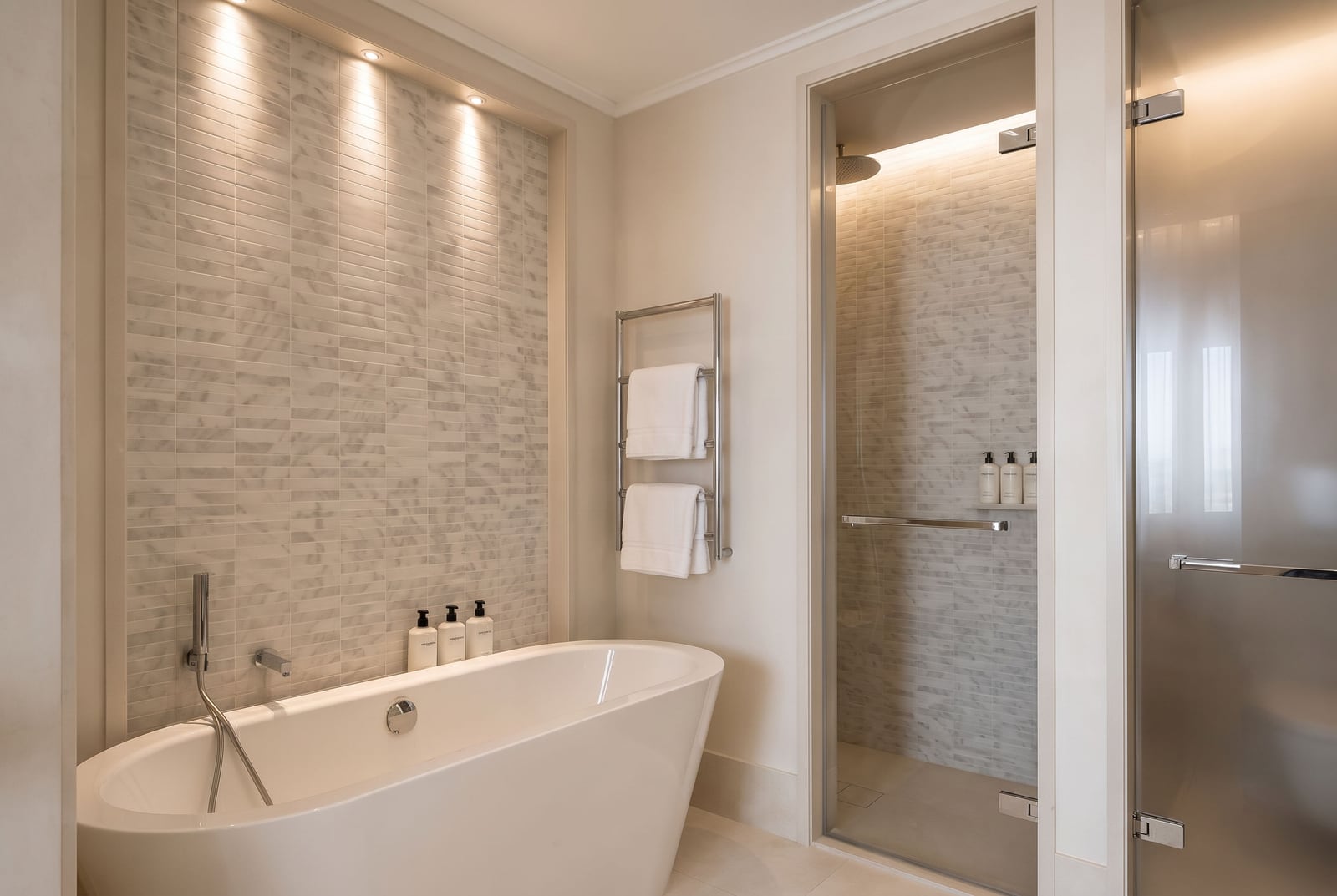 Modern bathroom featuring a freestanding bathtub, glass shower enclosure, heated towel rack, and marble tile walls with recessed lighting.