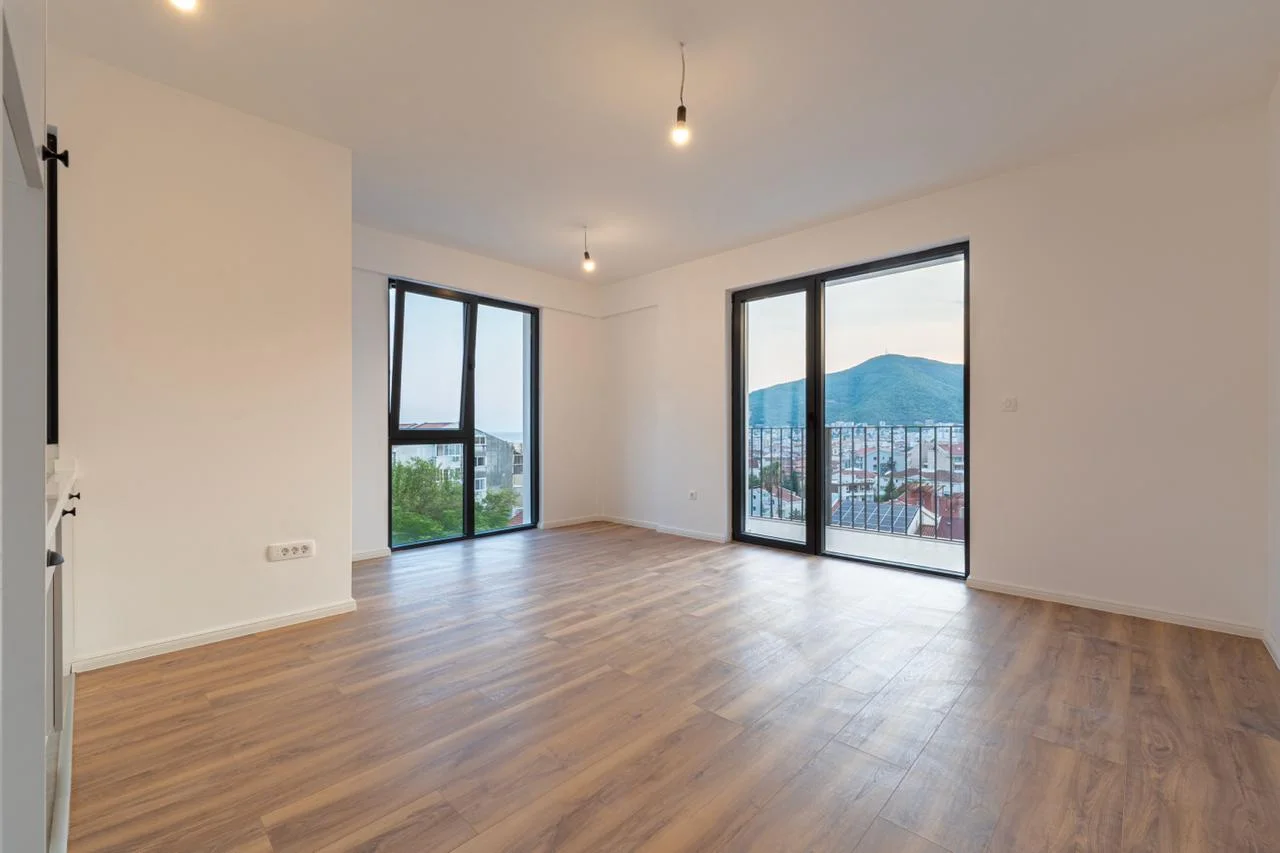 Empty room with wood flooring, large windows, and a balcony door overlooking mountains and city.
