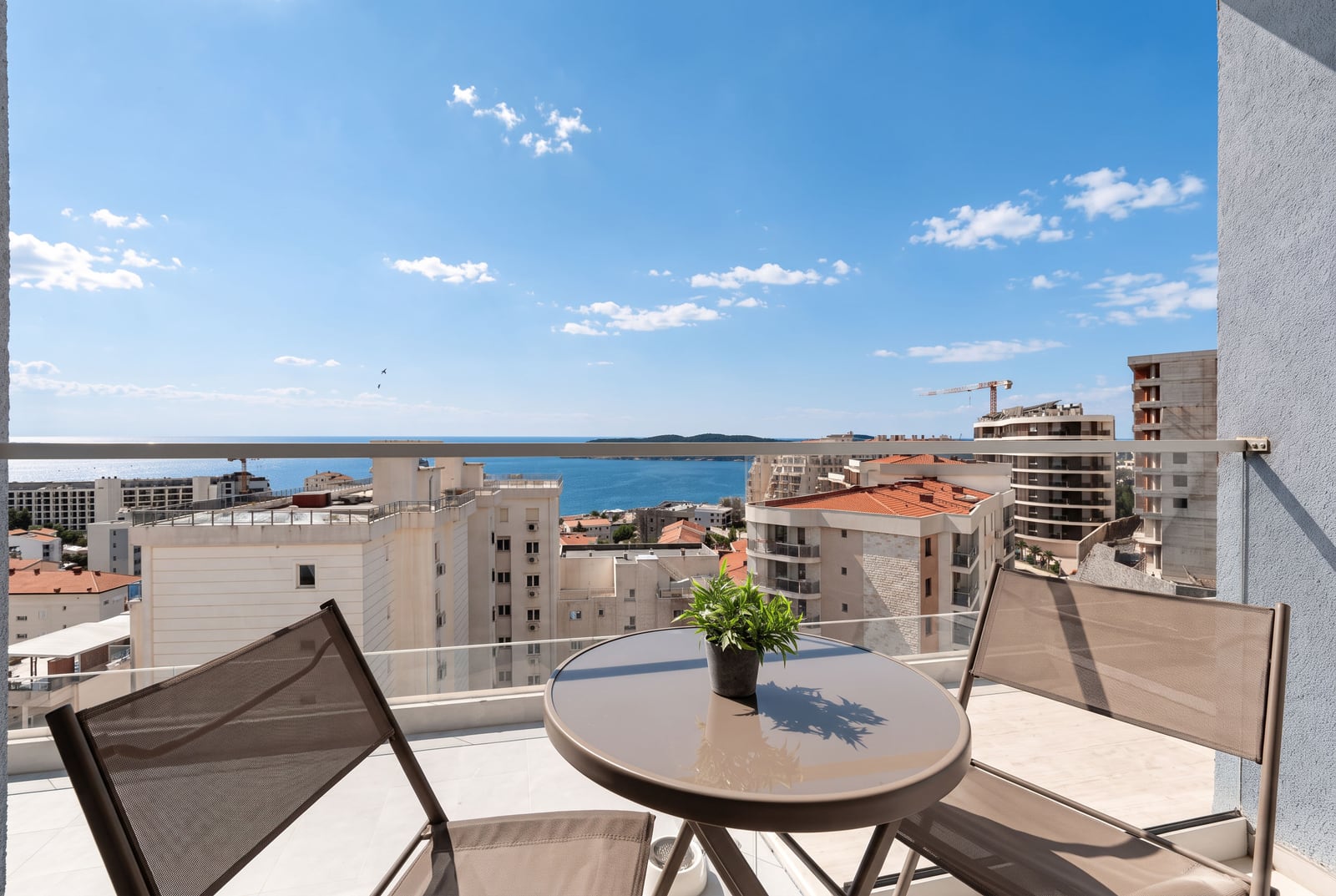 Balcony with sea and city view, featuring a small table with a plant and two chairs behind glass railing.