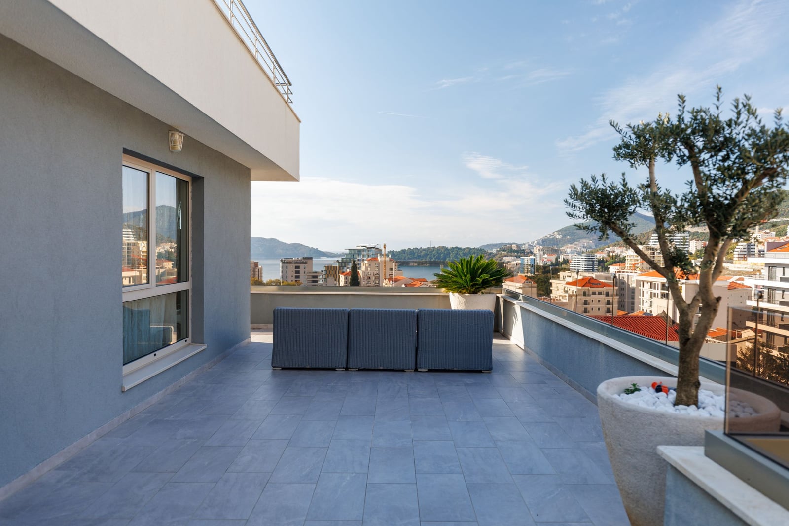 Spacious penthouse terrace in Becici, Montenegro, featuring wicker furniture, an olive tree, and panoramic sea views.