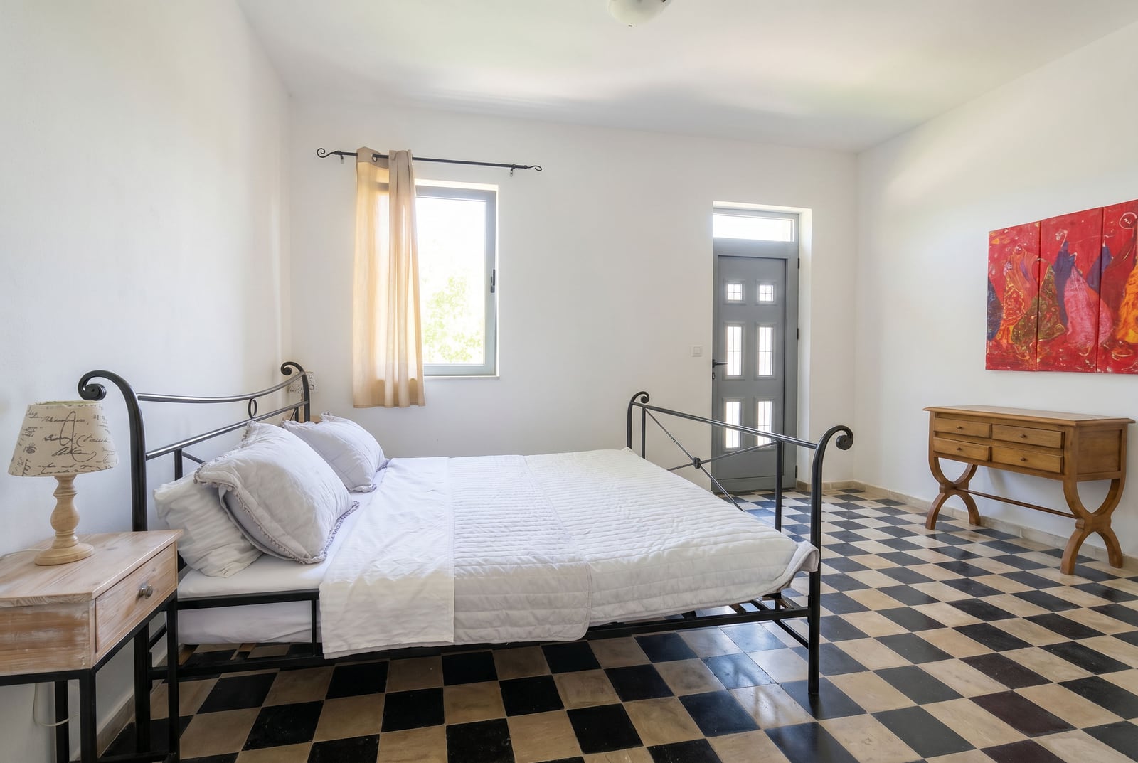 Bedroom in an apartment in Risan, Montenegro: wrought iron bed, checkerboard floor, wooden console, and a bright painting on the wall.