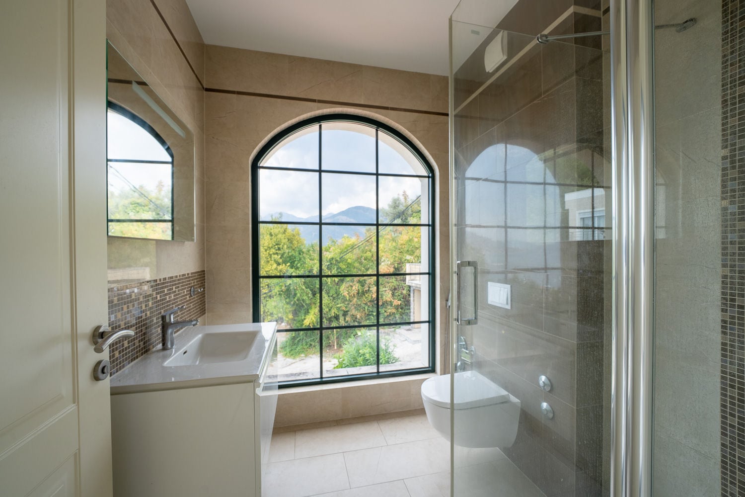 Bathroom with glass shower enclosure, vanity sink, wall-mounted toilet, and arched window overlooking mountains.