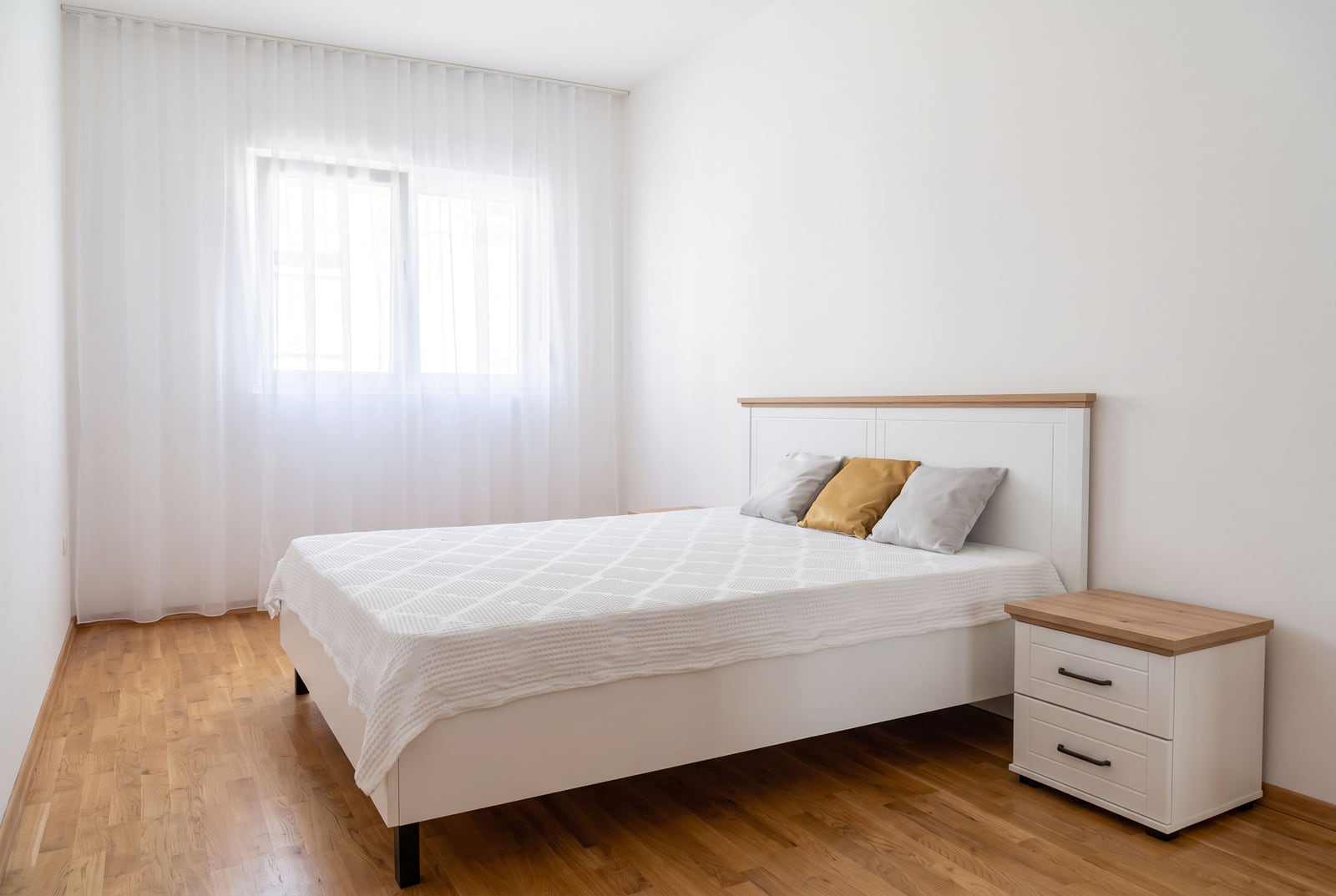 Bright bedroom featuring a white bed, wooden nightstand, parquet flooring, and a window with sheer curtains.