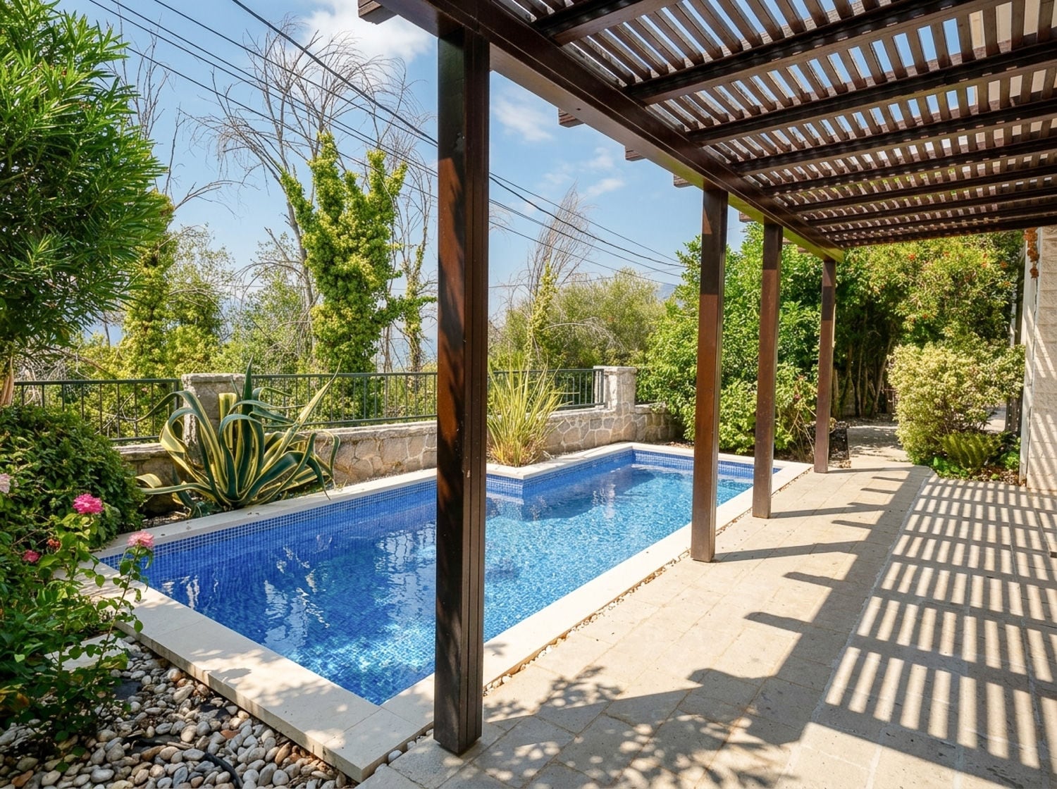 A blue swimming pool is visible under a wooden pergola surrounded by lush greenery and stone paving.