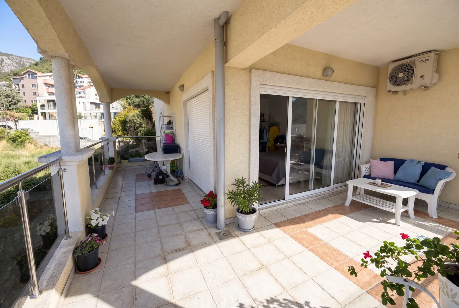 Long tiled balcony with white plastic furniture, potted flowers, glass railings, and a sliding door leading to an interior room.