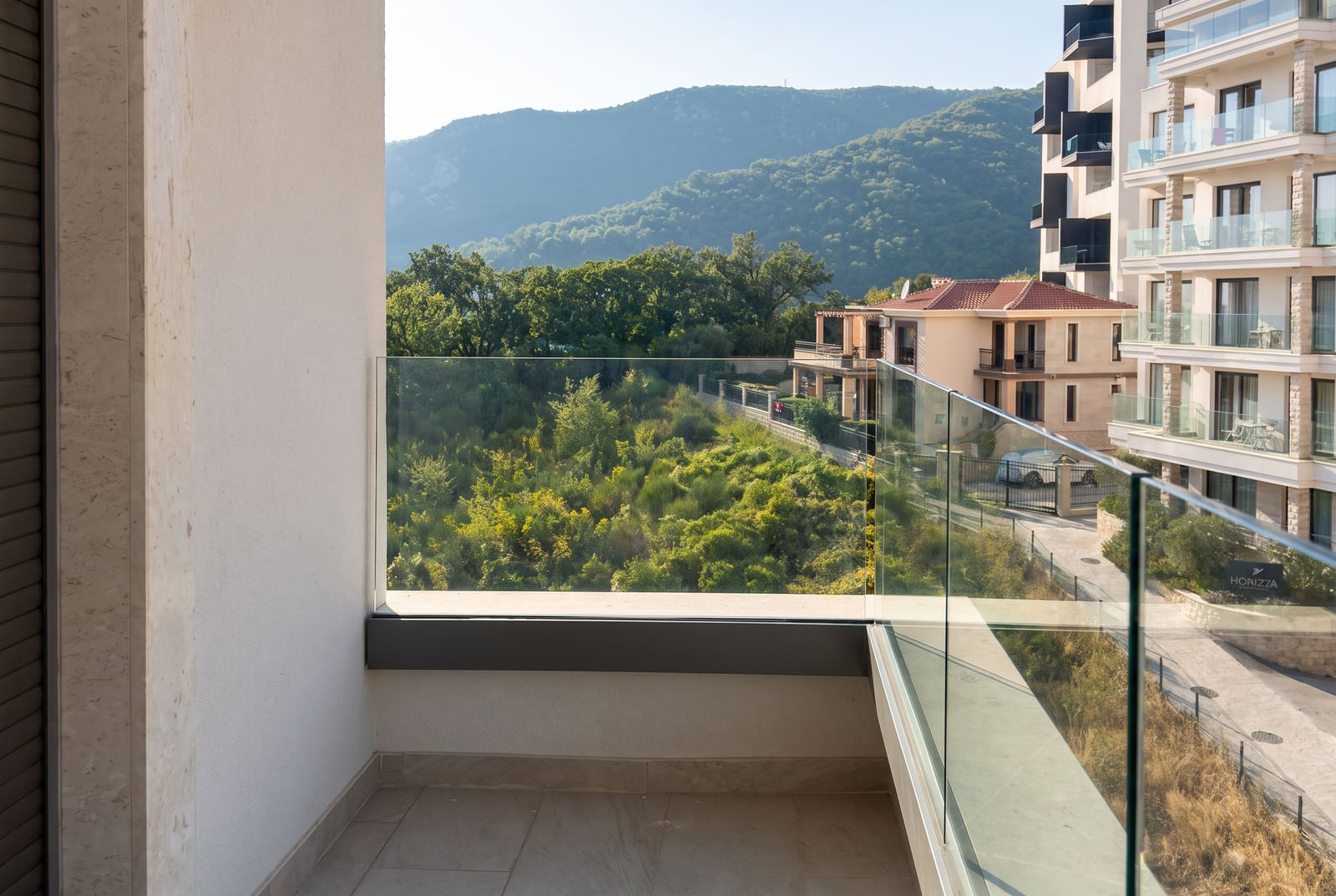 Balcony with glass railing overlooking green vegetation and mountains. Tiled floor and white wall visible.