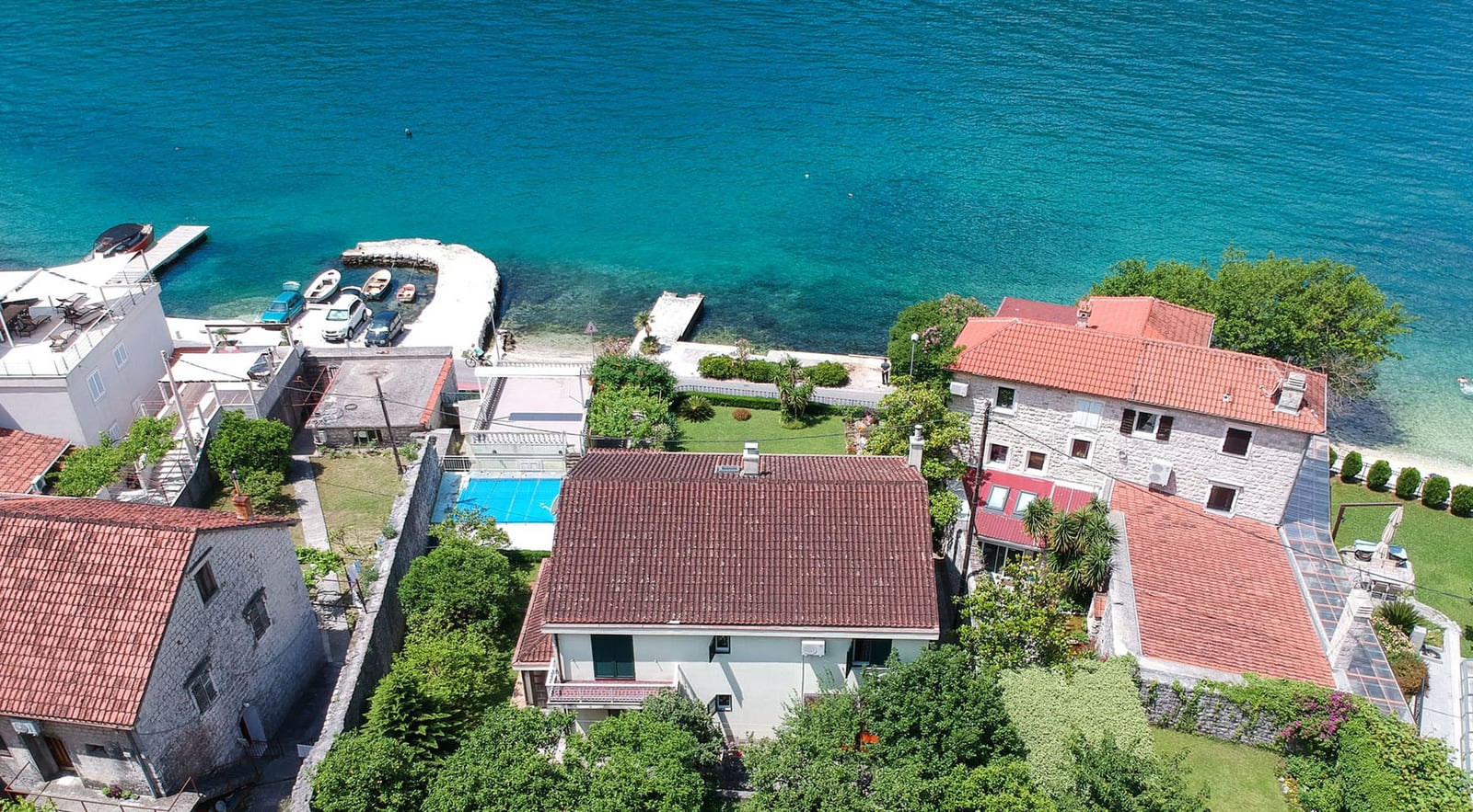 Aerial view of a villa in Dobrota, Montenegro: stone houses, a pool, a green garden, and direct access to the sea with a pier.