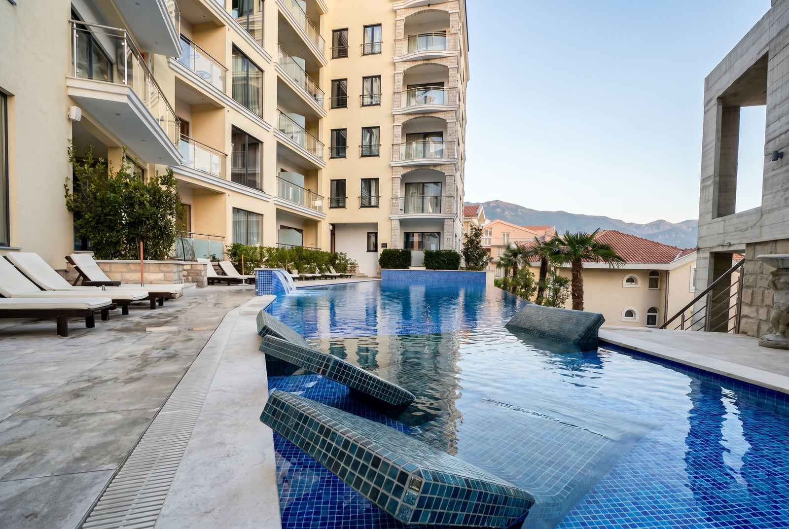 Outdoor swimming pool with sun loungers at a modern residential complex in Bečići, Montenegro, with a view of the mountains and terrace.