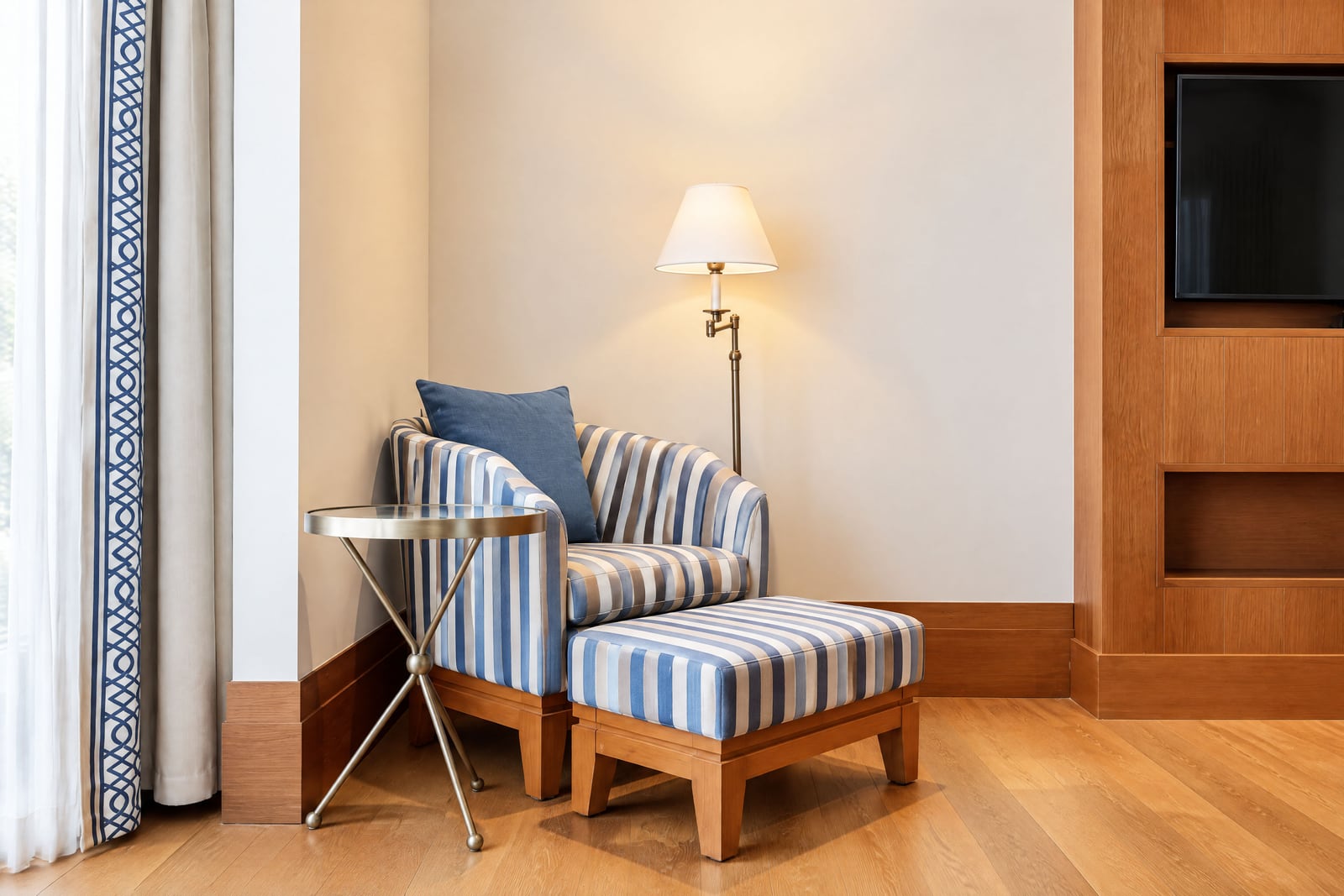 Corner seating area with striped armchair, ottoman, floor lamp, and wooden TV unit.