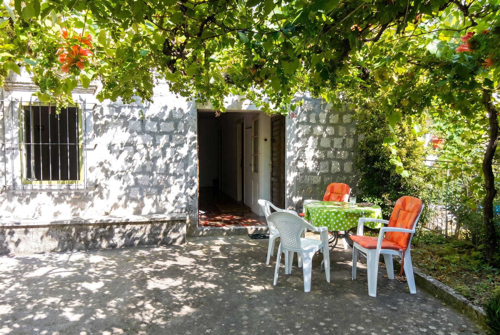 Cozy terrace of a stone house in Dobrota, Montenegro, featuring a table, chairs, and a shaded grapevine in the courtyard.