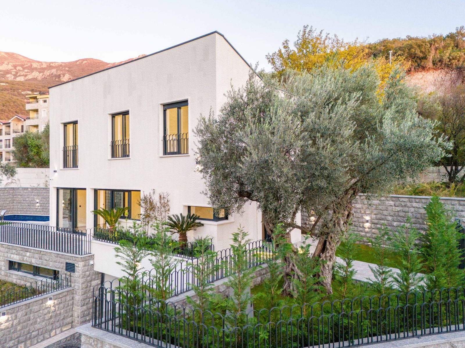 Modern white villa in Kamenovo, Montenegro: facade with panoramic windows, swimming pool, garden with olive tree and fence.