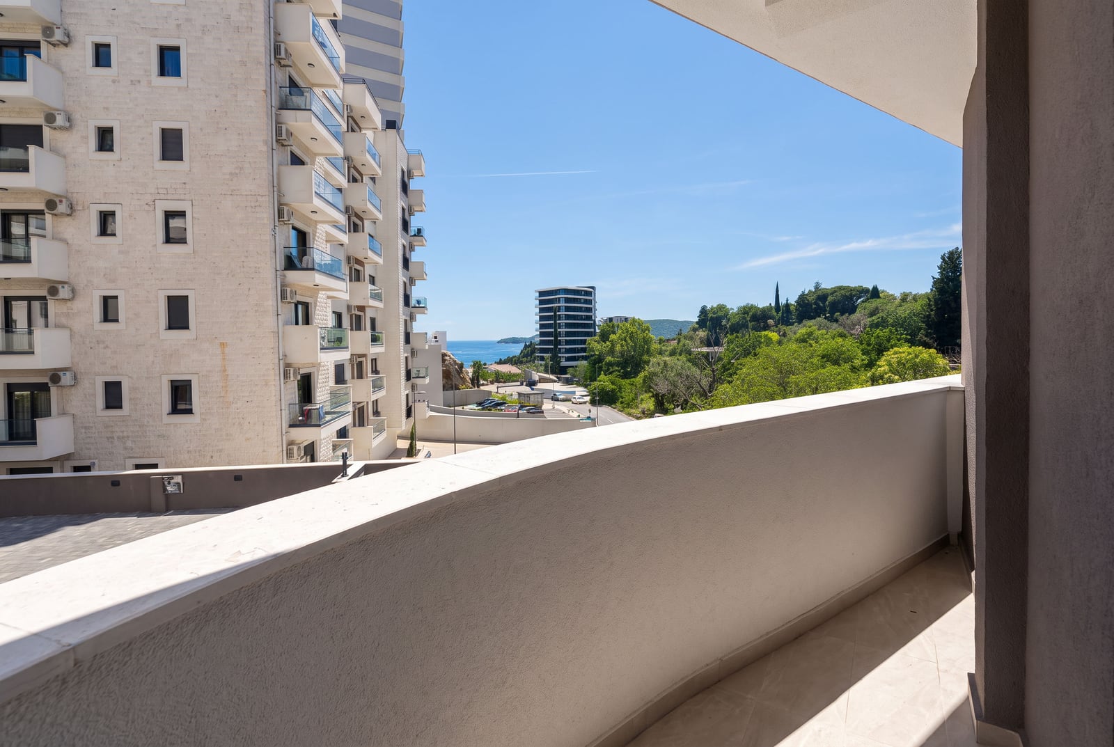 View from a tiled balcony showing a neighboring apartment building, green trees, and the sea in the distance.