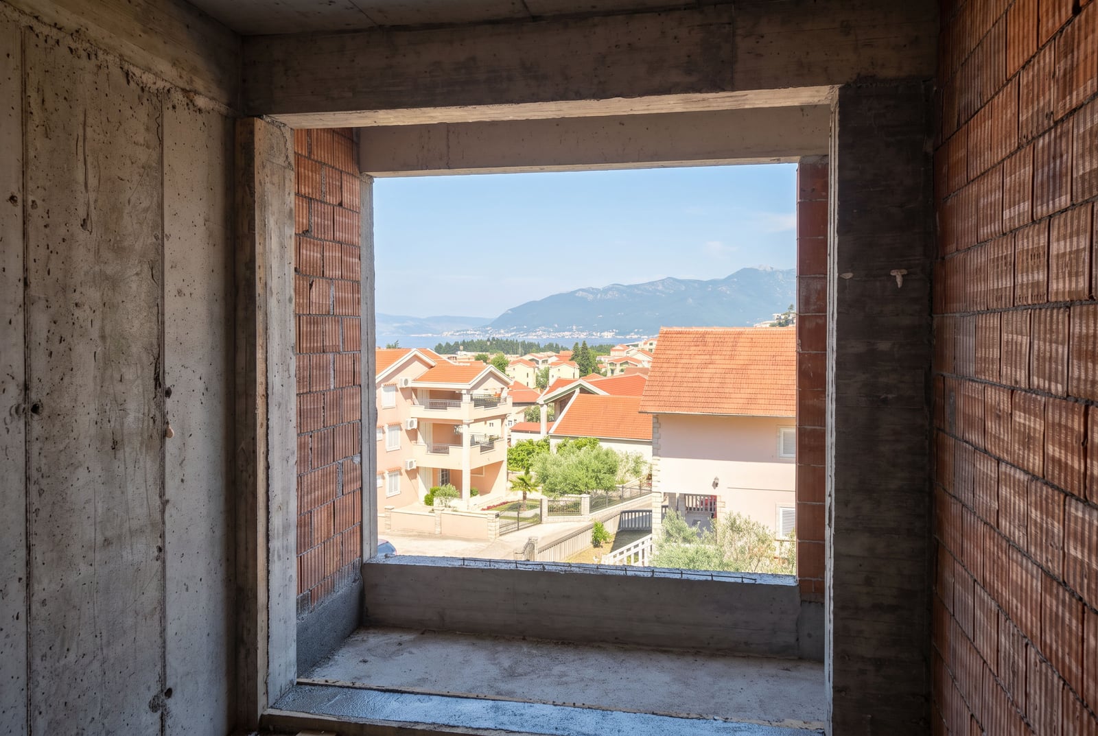 Unfinished construction interior showing a window opening with brick and concrete framing a view of houses and mountains.