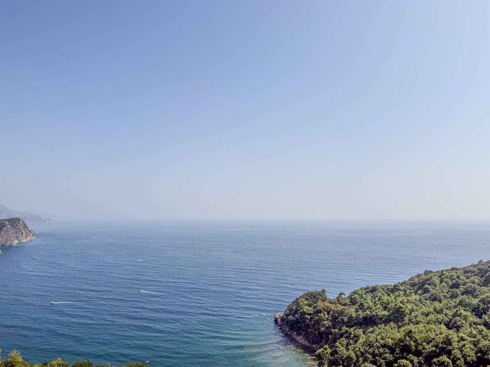 Panoramic view of the sea, forested coastline, and clear sky. Rocky cliffs and boats visible.
