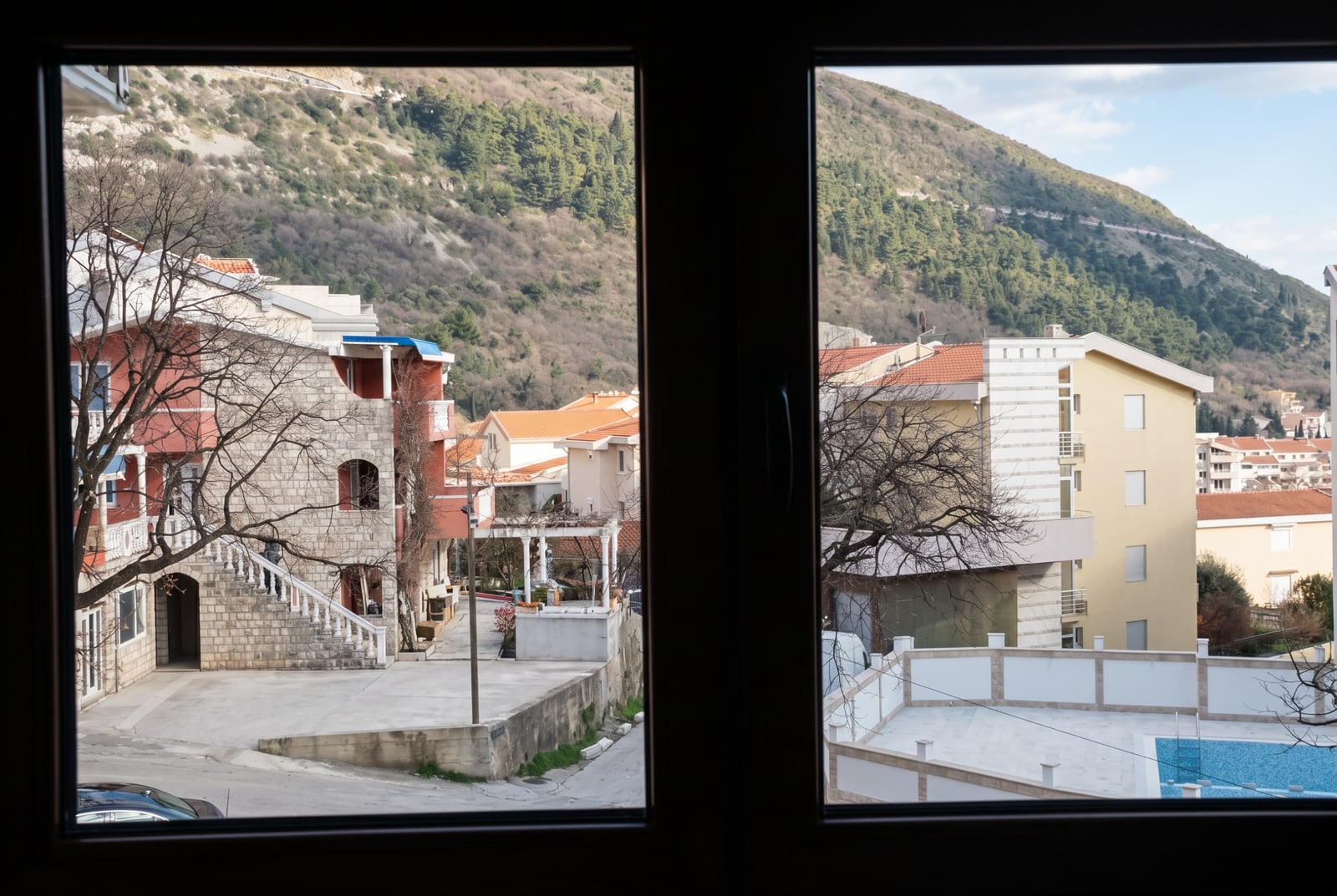 View from the apartment window in Budva, Montenegro: panoramic view of the mountains, residential buildings with tiled roofs, and an outdoor swimming pool.