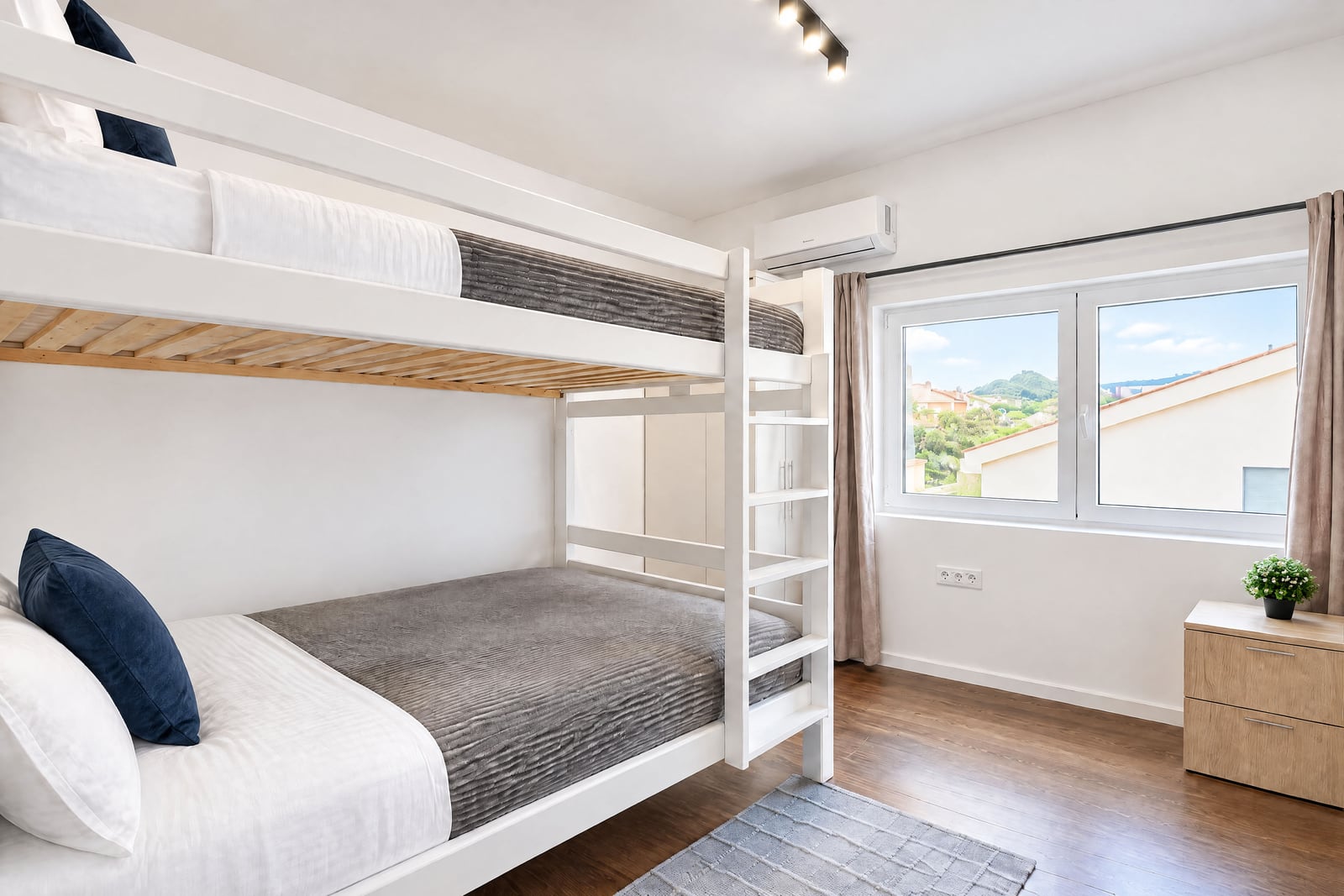 White bunk bed with gray bedding, wooden floor, window showing rooftops, and a wall-mounted air conditioner unit.