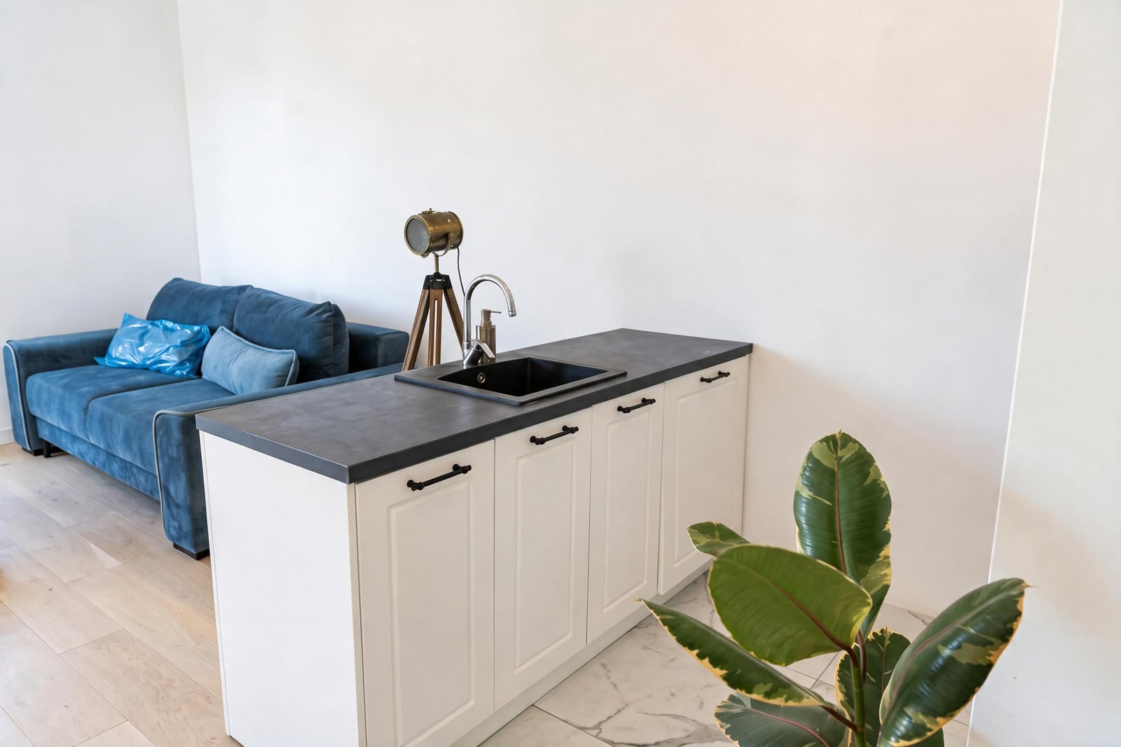 White kitchen with a dark countertop and sink, blue sofa and a houseplant in the living room of an apartment in Tivat, Montenegro.