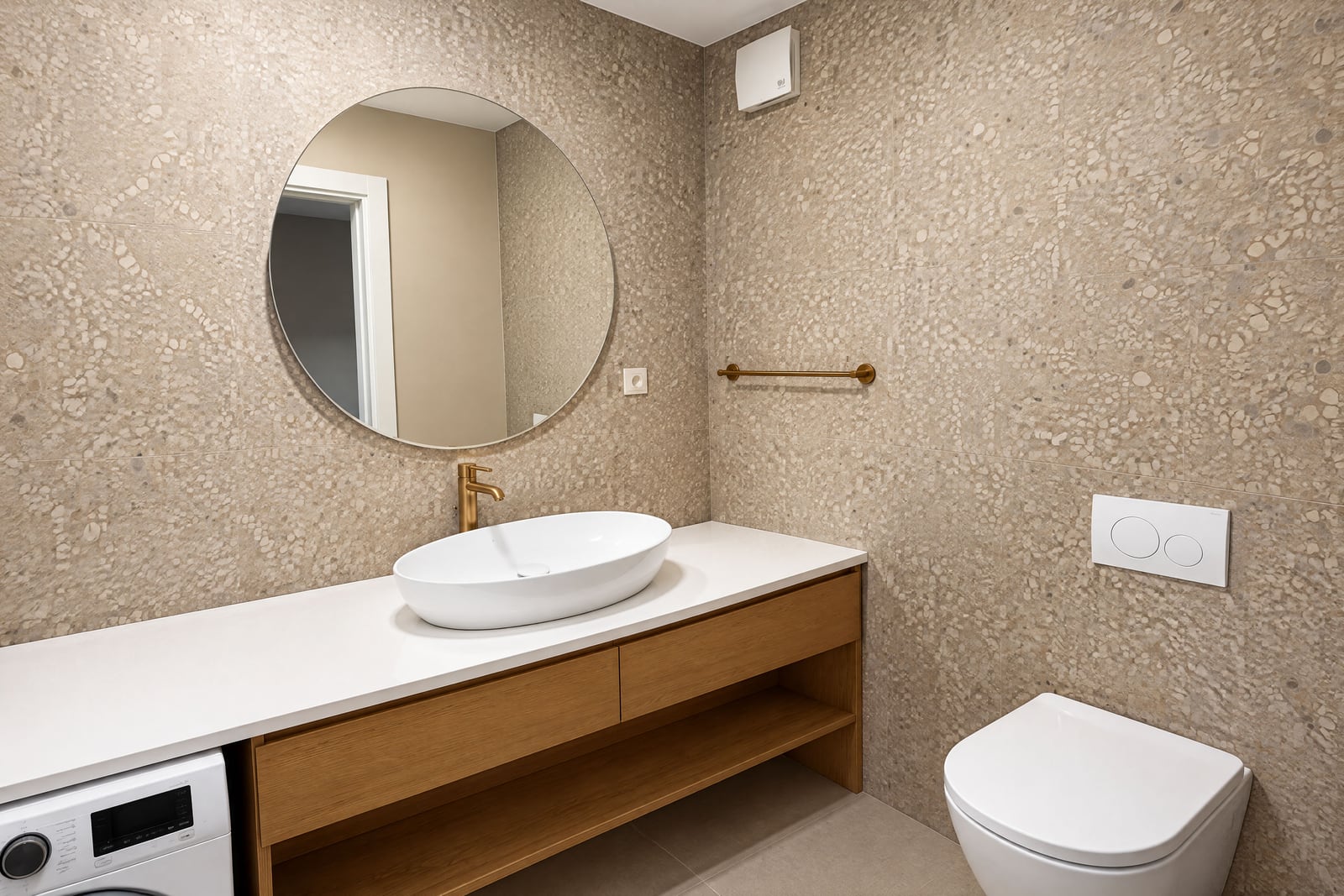 Bathroom with terrazzo tiles, round mirror, wooden vanity, vessel sink, and wall-mounted toilet.