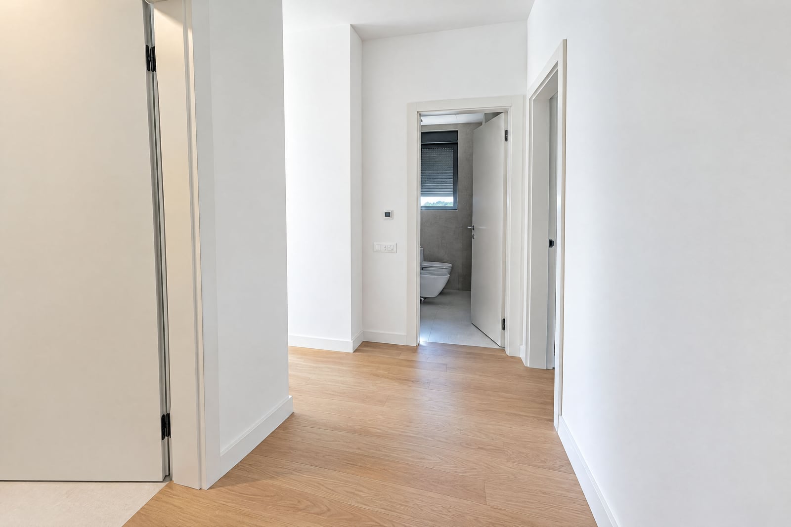 White hallway with light wood flooring and an open door leading to a bathroom with a visible toilet.