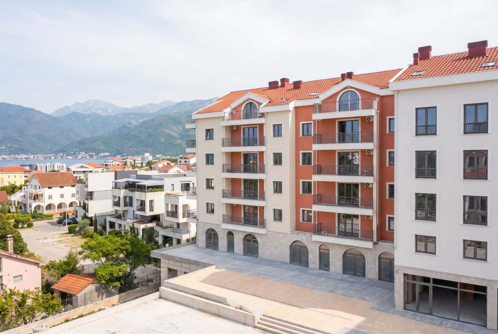 View of a multi-story residential complex with terracotta roof and balconies against mountains and sea.