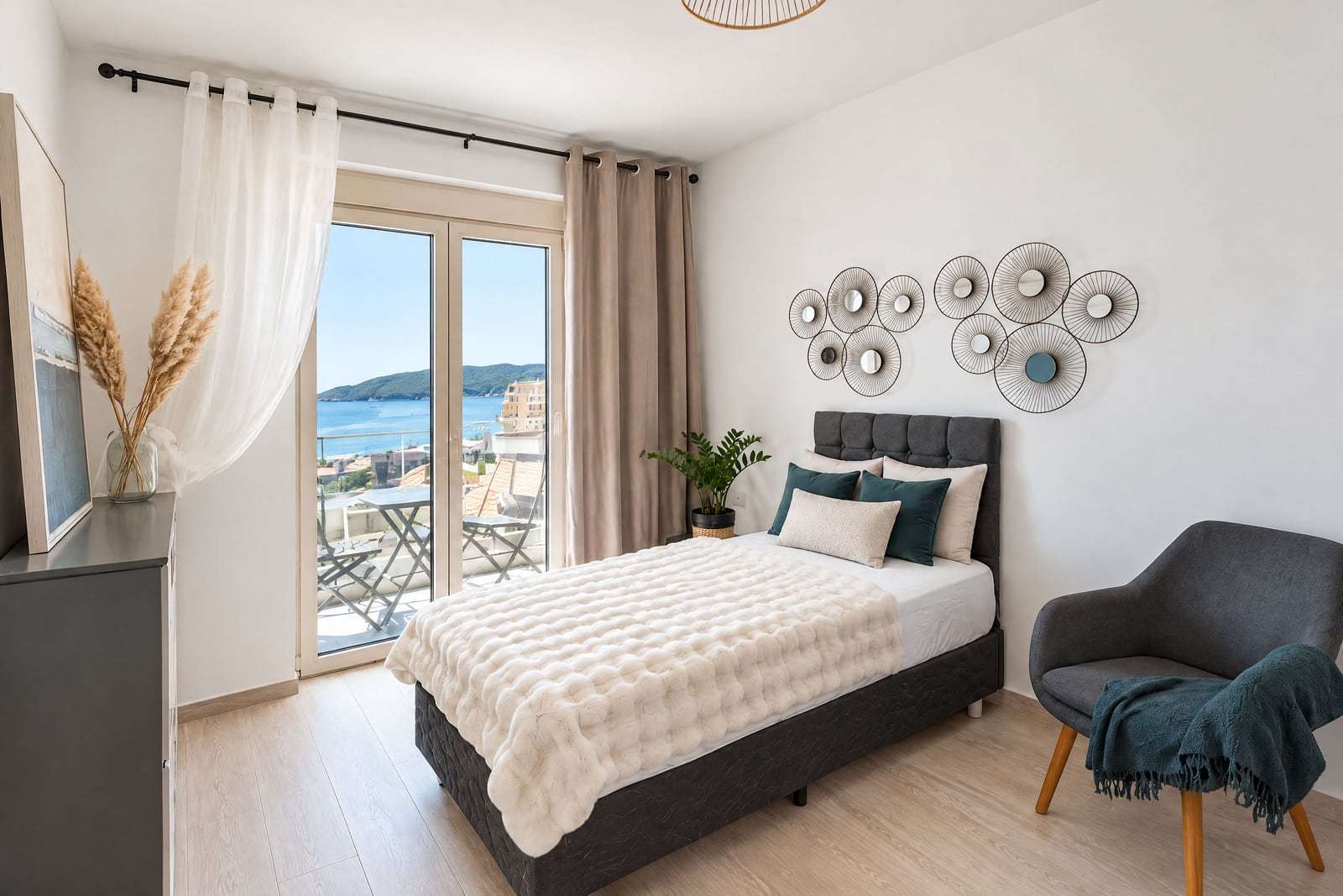 Modern bedroom featuring a double bed, grey armchair, and balcony door overlooking the sea and hills.