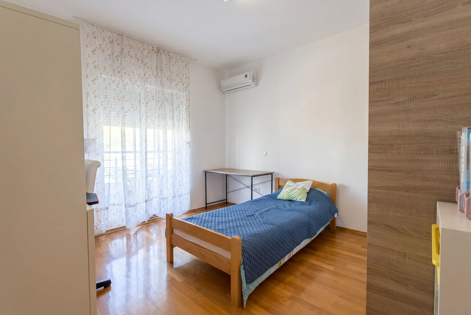 Bright bedroom featuring a single bed with blue cover, wooden desk, wall-mounted AC unit, and sheer curtains.