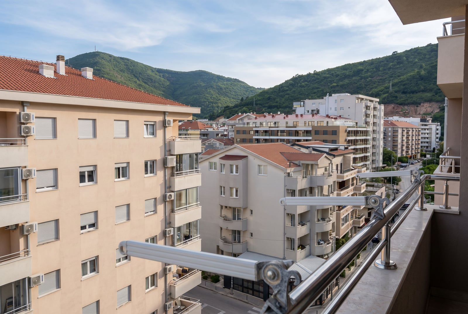 View from a balcony showing nearby apartment buildings, a street, and green mountains in the background.