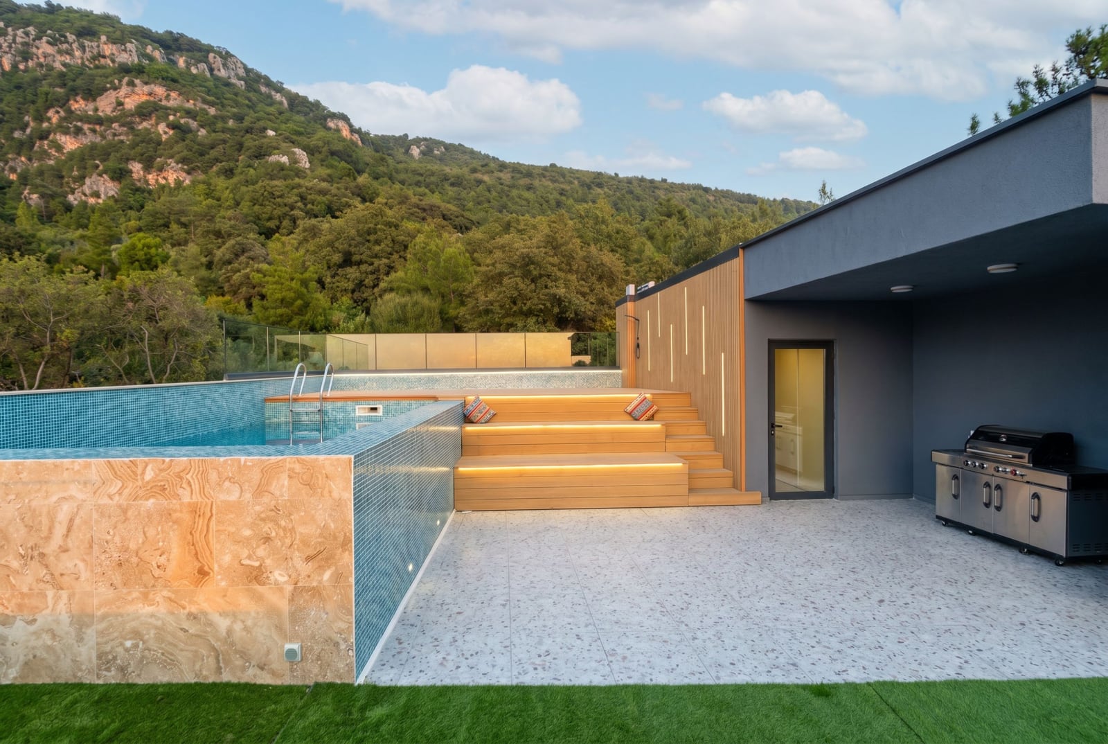 Villa terrace in Kavac, Montenegro: illuminated pool, wooden lounge area, grill, and mountain view.