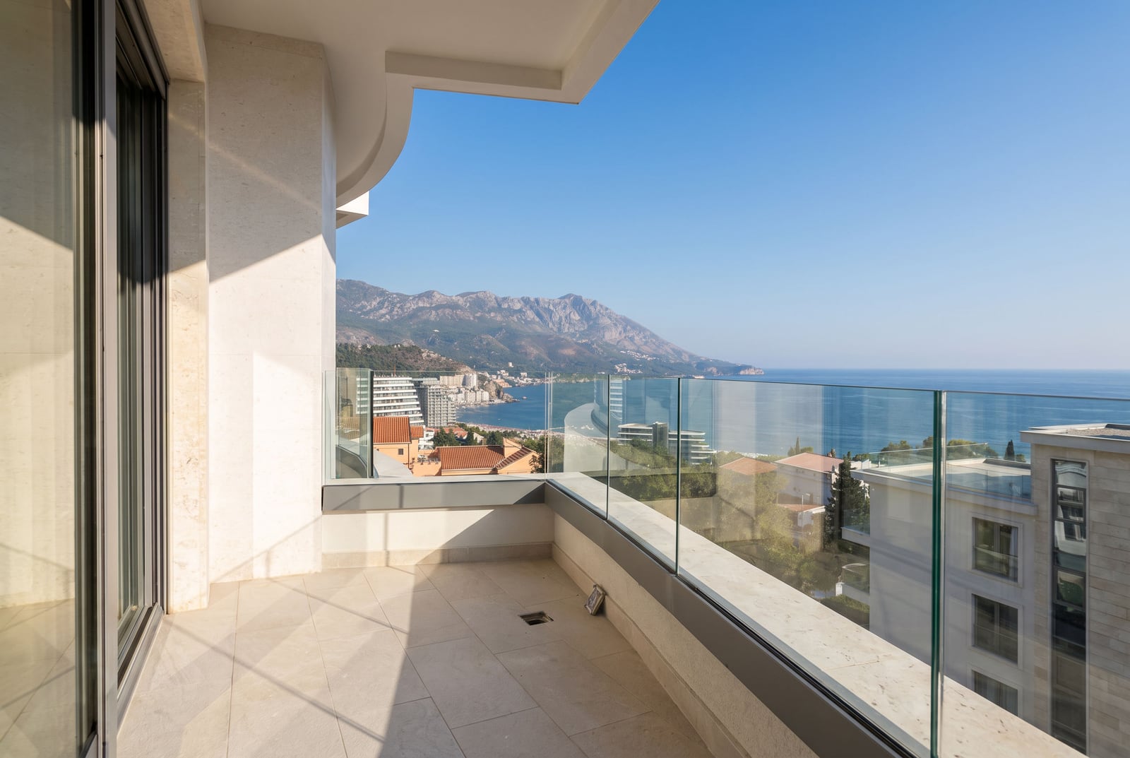 Modern balcony with glass railing offering a panoramic view of the sea and mountains.
