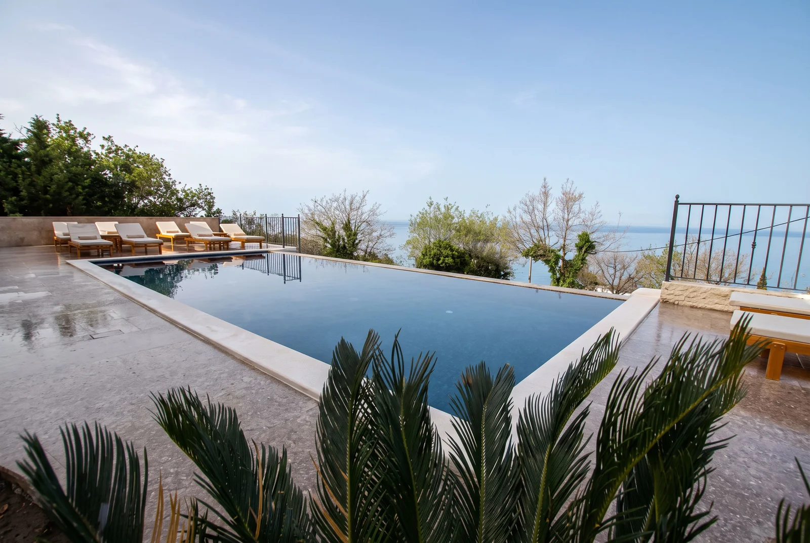 Infinity pool with lounge chairs overlooking the sea and greenery under a clear sky.